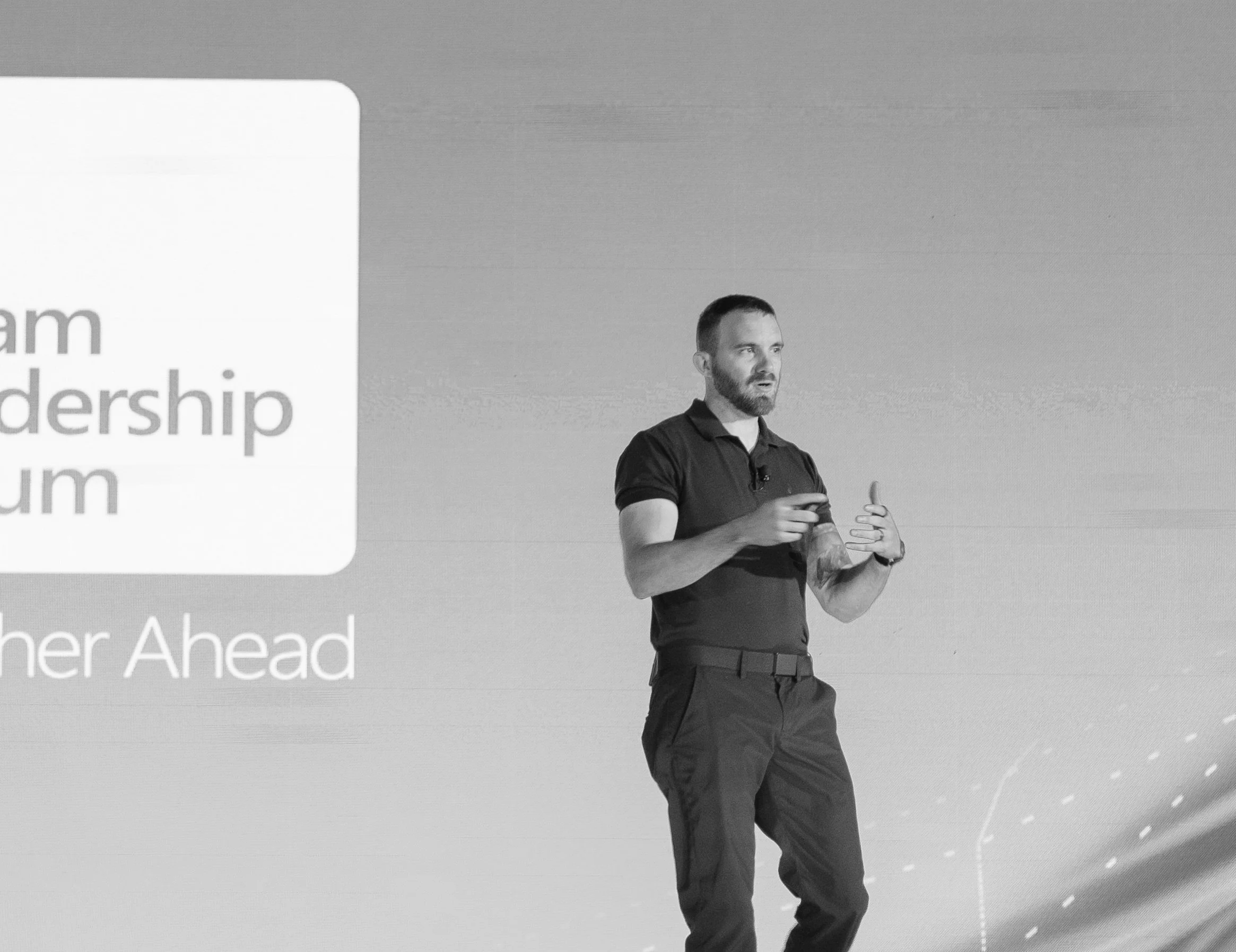 A man is standing on a stage giving a presentation. There is a projected slide behind him with partially visible text that reads 'Leadership' and 'Ahead.' The man has a beard, short hair, and is wearing a dark polo shirt and dark pants.