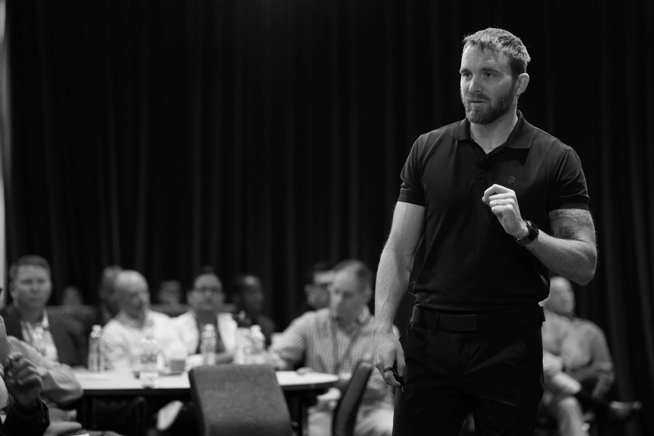 A man in a black polo shirt speaking at a conference, with an audience seated at tables watching him.
