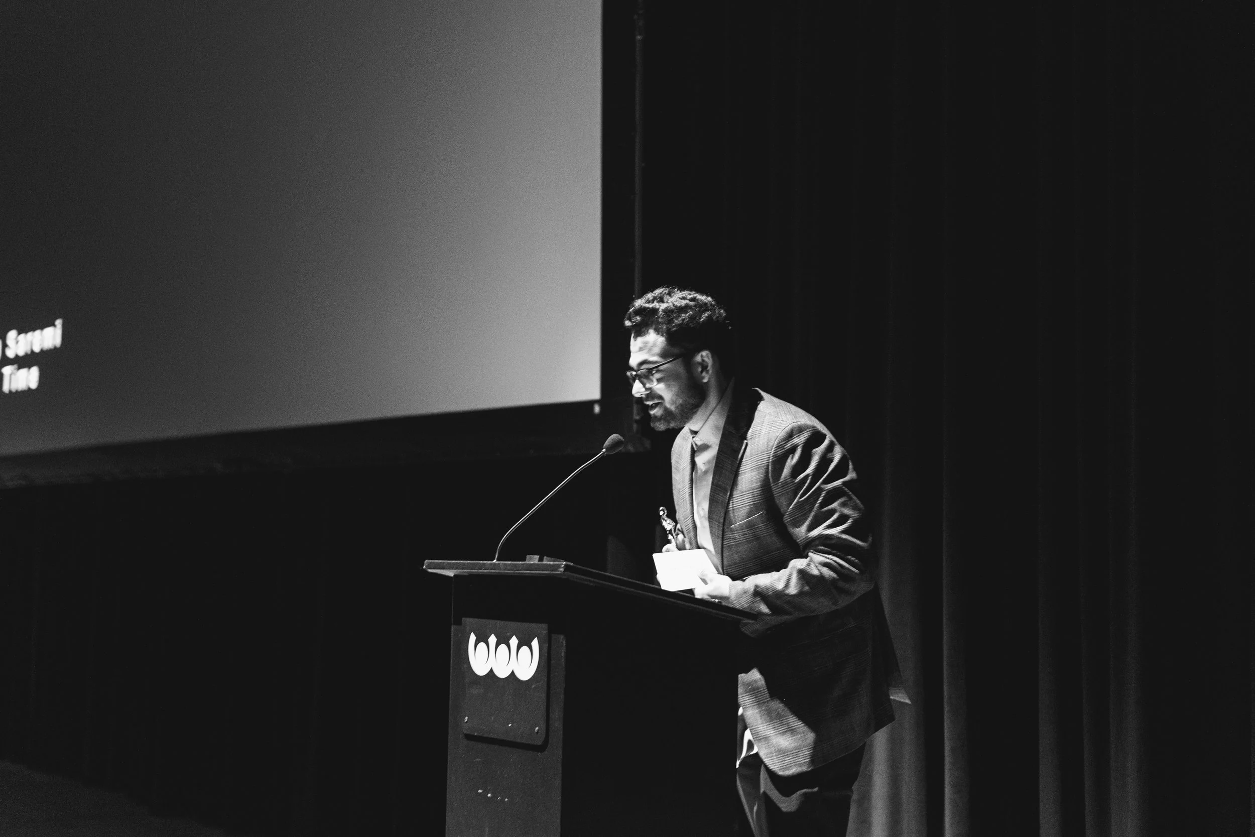 A man with glasses and a beard in a checkered blazer speaking at a podium on a stage in a black and white photo.