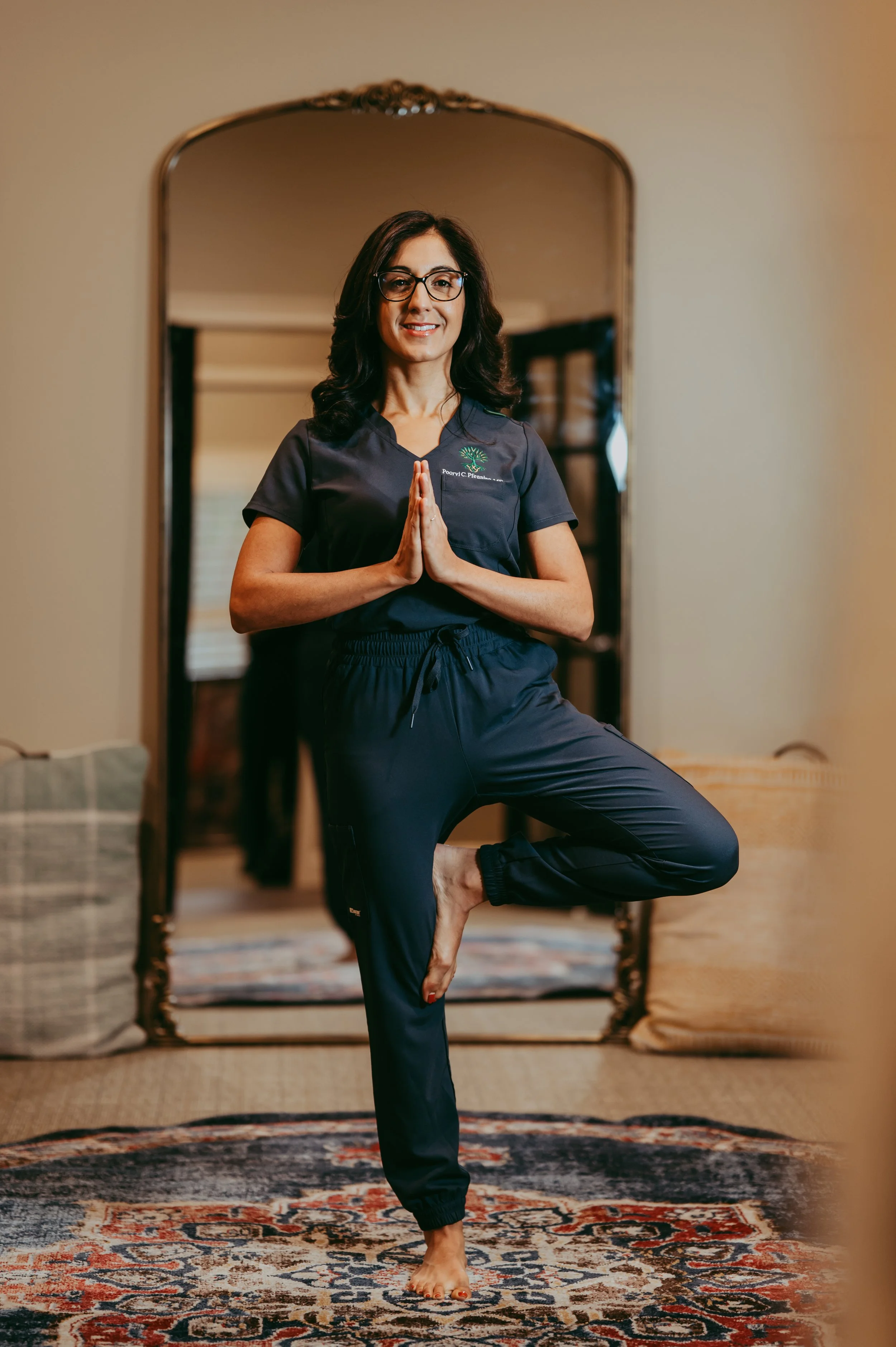 A woman practicing yoga in front of a mirror in a living room. She is standing on one leg, with her hands in a prayer position at her chest, and is smiling. She wears glasses and a dark workout outfit.
