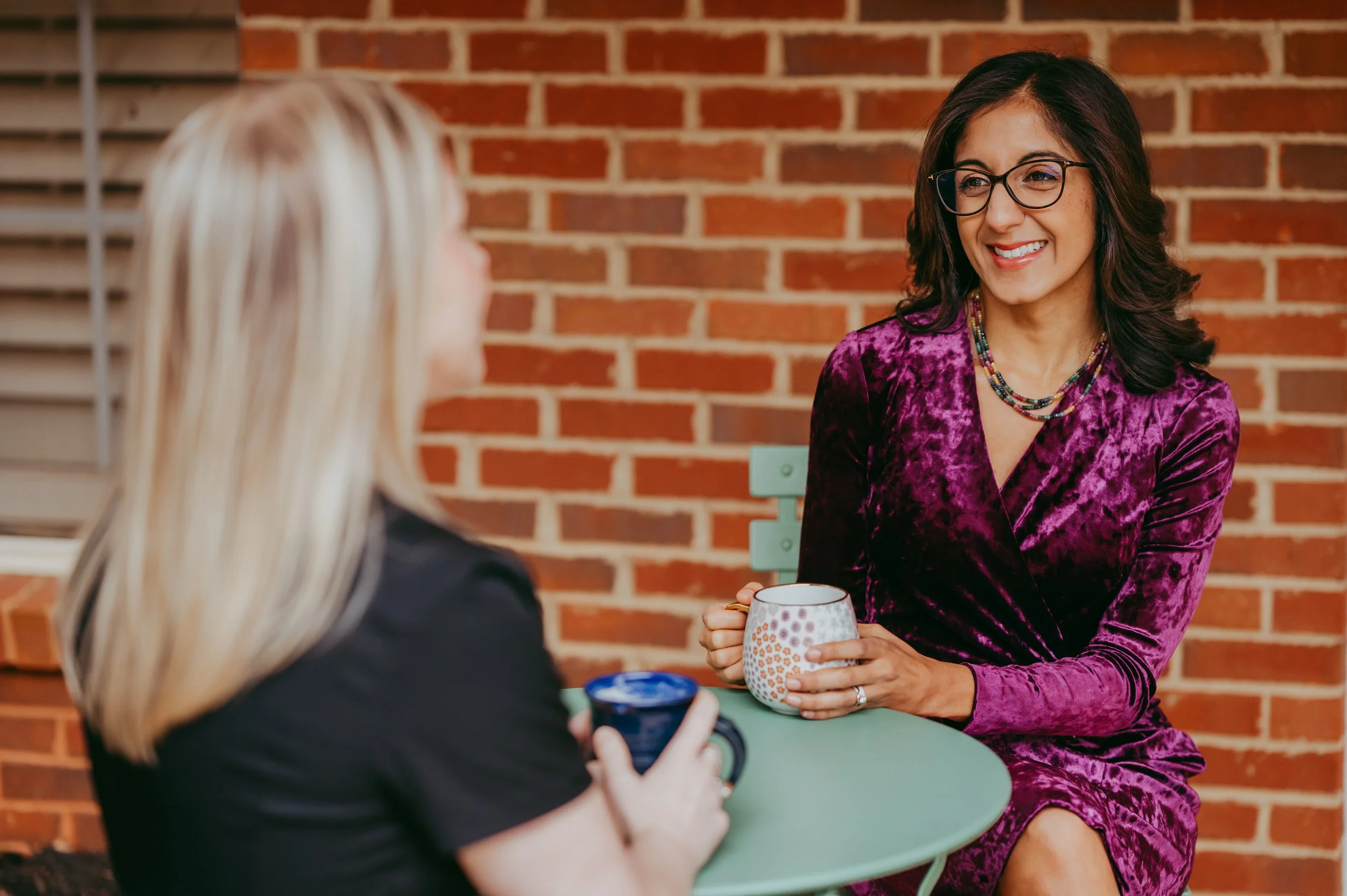 Two women sitting at a small round table outdoors, enjoying coffee or tea, with a brick wall in the background. One woman with blonde hair is blurred and facing away. The other woman has dark hair, glasses, and is smiling, wearing a purple velvet dress and layered necklaces.
