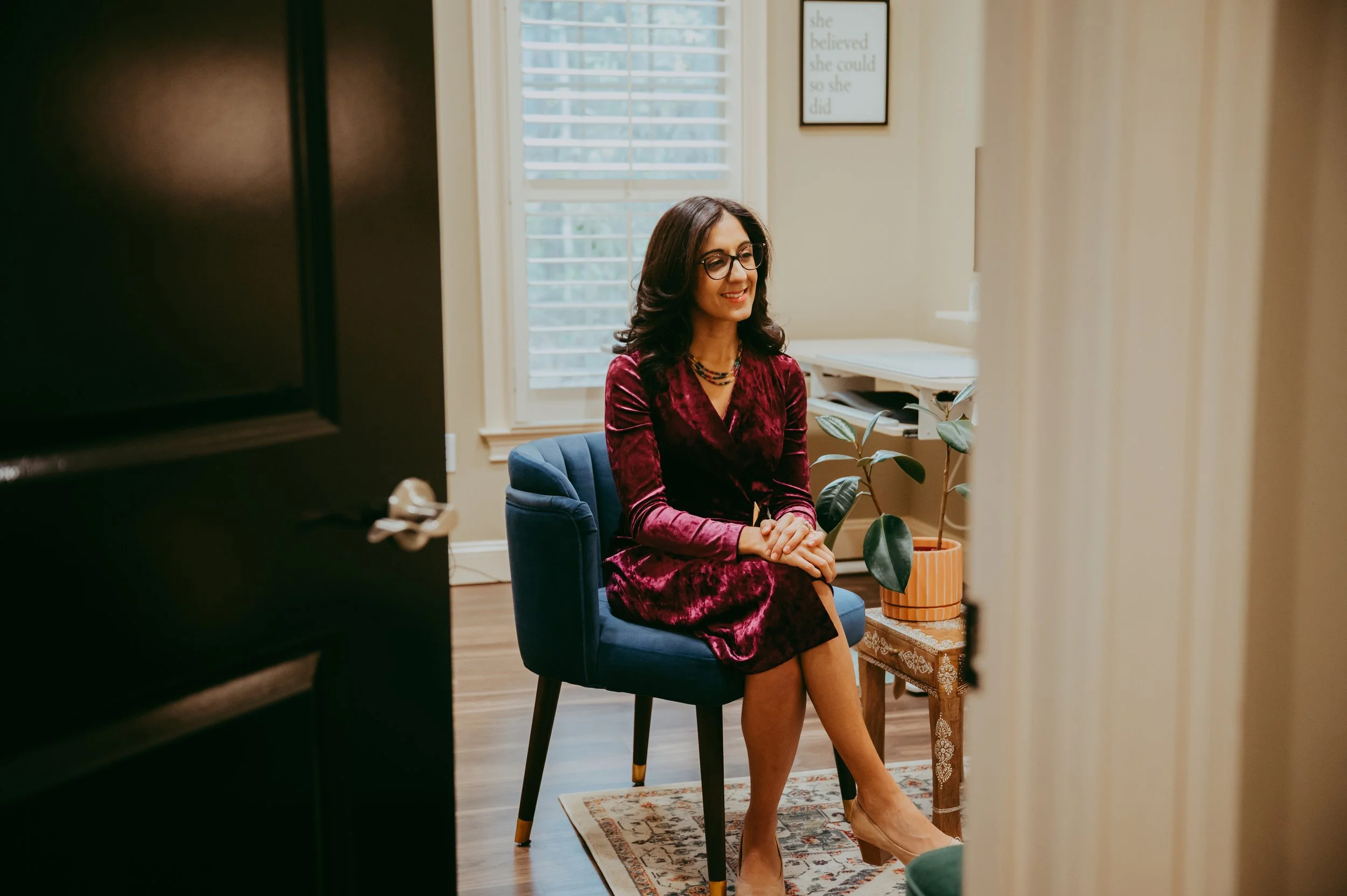 A woman with glasses and dark wavy hair, wearing a red velvet dress, sits on a blue chair in a cozy, brightly lit room. She's smiling, with a potted plant on a small wooden table beside her, and framed quotes on the wall behind her.
