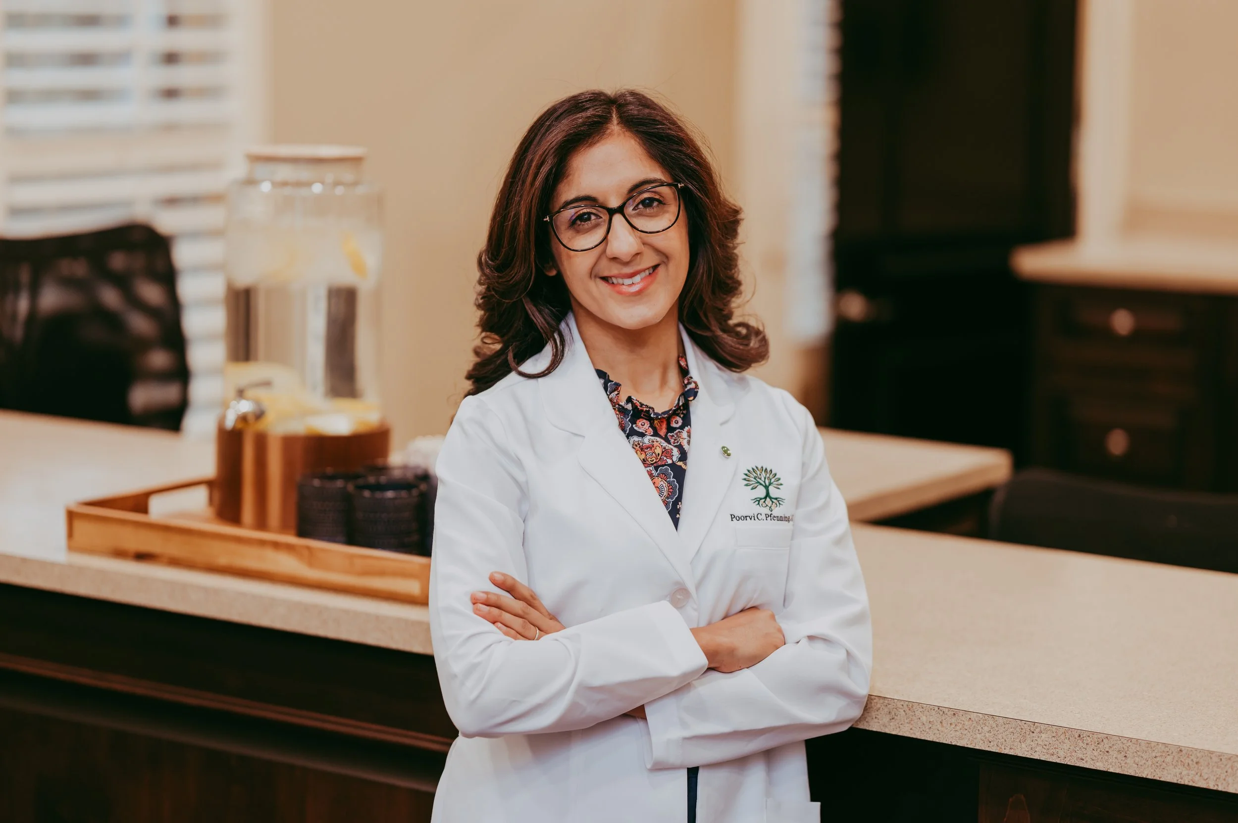 A woman with brown hair, glasses, and a white lab coat standing behind a counter, smiling with arms crossed in a professional setting.