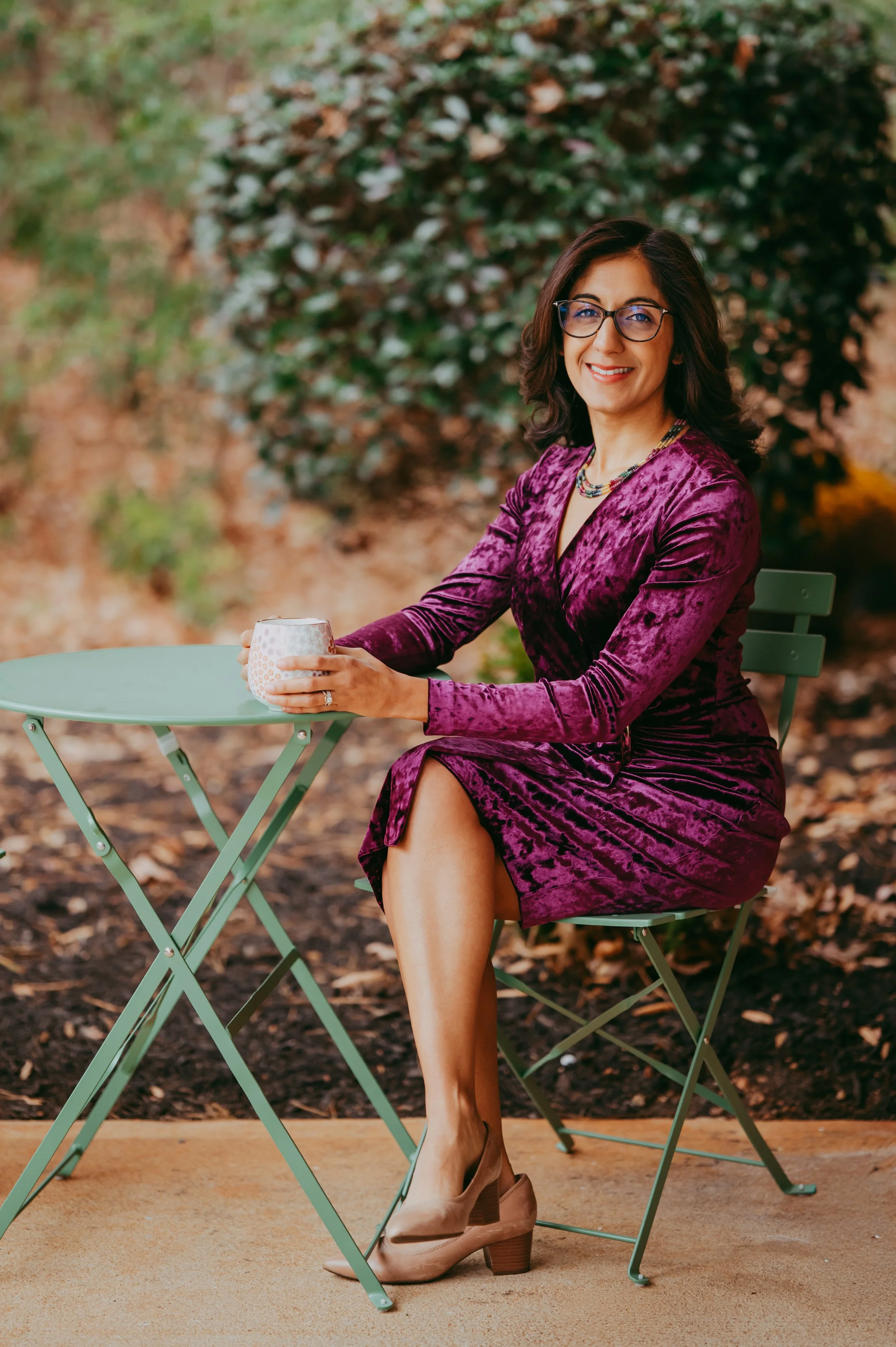 A woman with dark hair, glasses, wearing a purple velvet dress and beige heels, sitting at a green metal table outdoors, holding a mug, smiling.