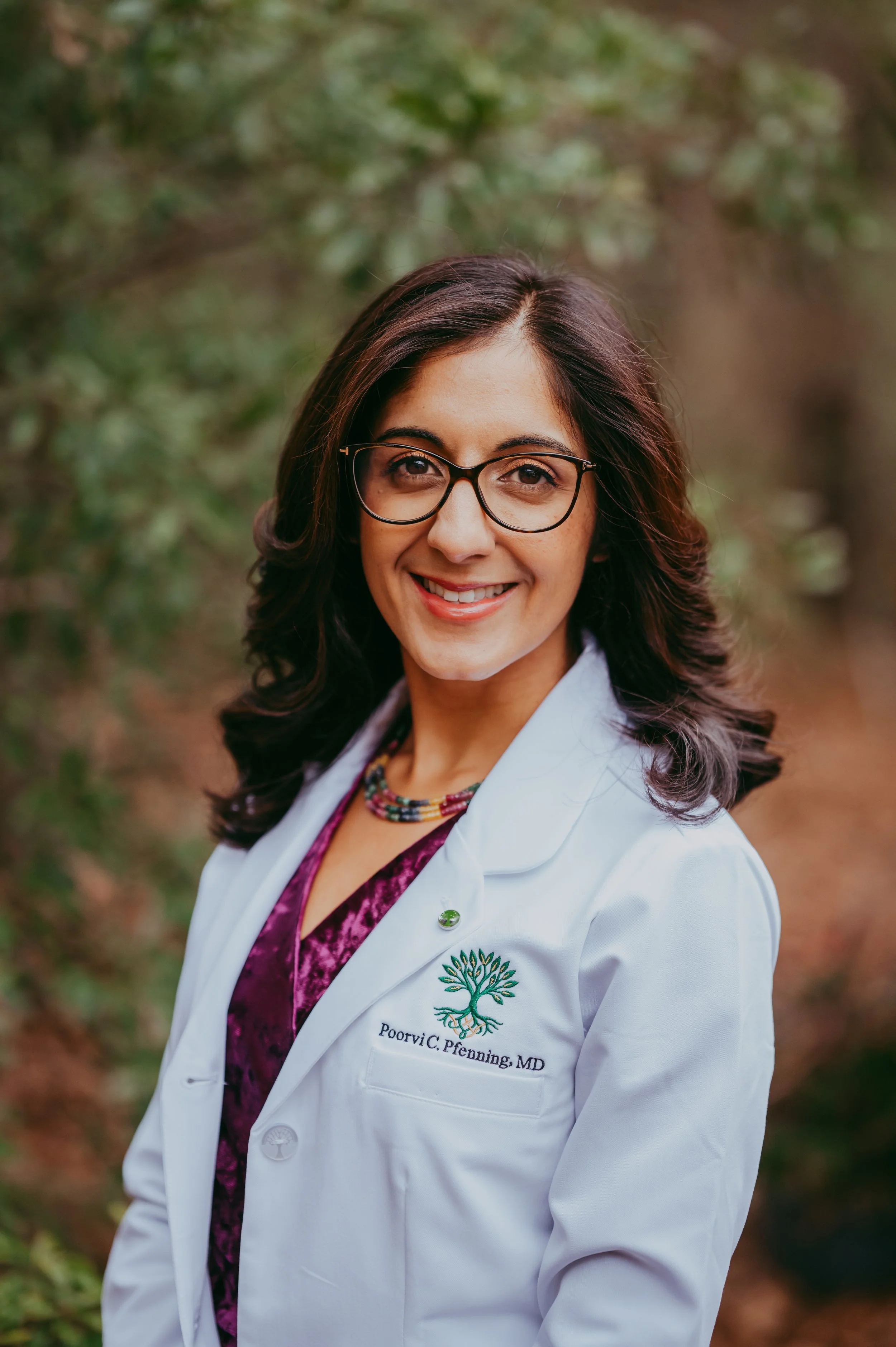 A woman with dark brown hair, glasses, and a white coat with a green tree logo and her name 'Poorvi C. Pfenning, MD' embroidered on it, standing outdoors with green foliage in the background, smiling.