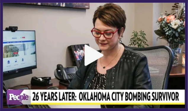 A woman with short hair and red glasses sitting at a desk in an office, with a computer monitor, phone, and flowers in the background, on a news segment about an Oklahoma City bombing survivor.
