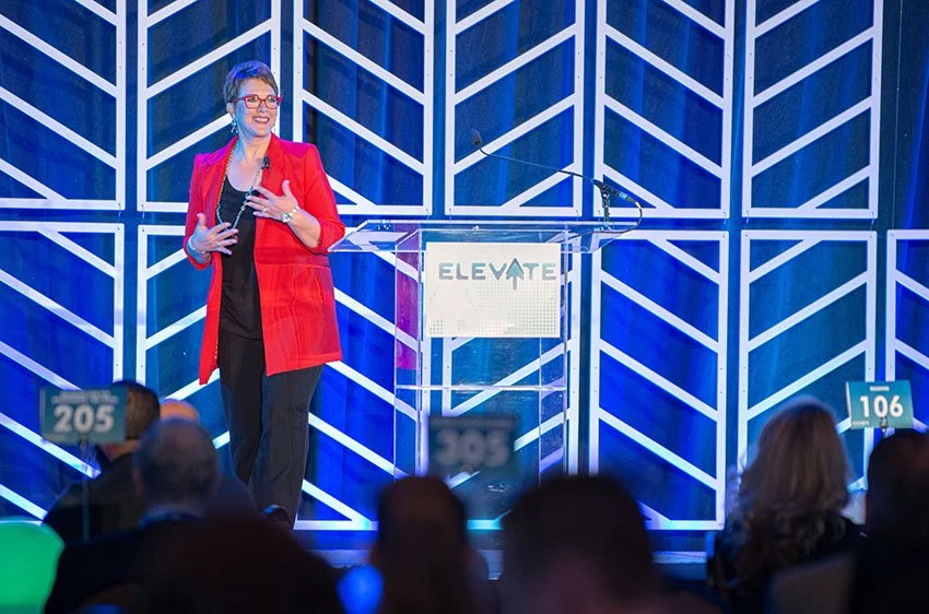 A woman in a red blazer and black outfit speaking at a conference stage with a clear podium that says 'ELEVATE'. She is standing in front of a geometric, blue-lit backdrop, and there are audience members in the foreground holding signs with seat numbers.