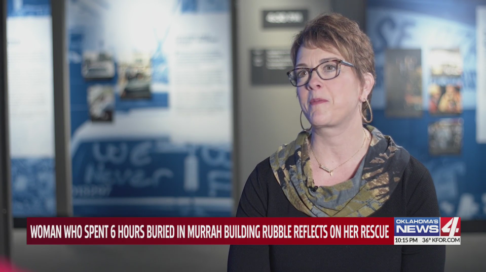 A woman with short hair and glasses being interviewed indoors, with a blurred informational display in the background. She is wearing a scarf and a dark top.