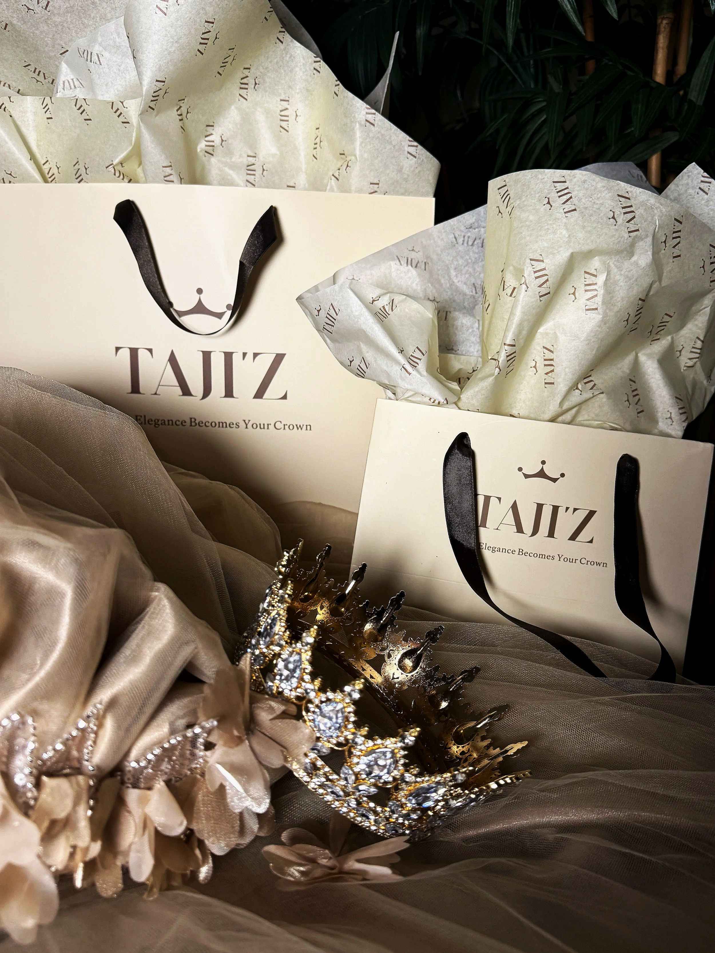Two elegant TAJ'Z shopping bags with black handles, filled with tissue paper, surrounded by a gold tiara with rhinestones and beige fabric with floral accents.