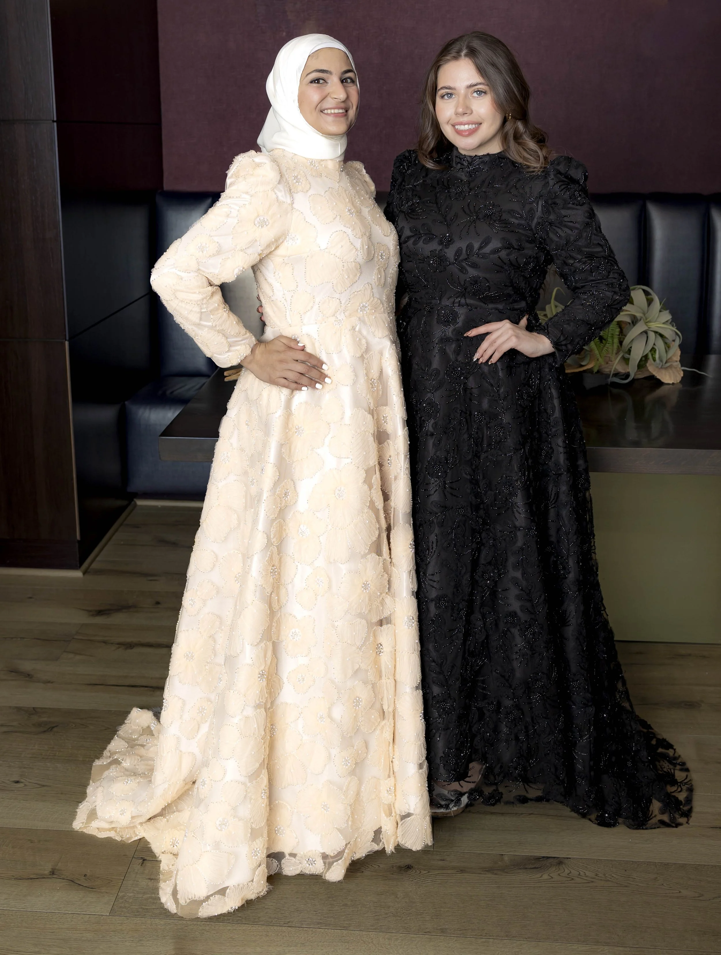 Two women in elegant dresses standing side by side, smiling, indoors with a dark purple wall and a bench behind them.