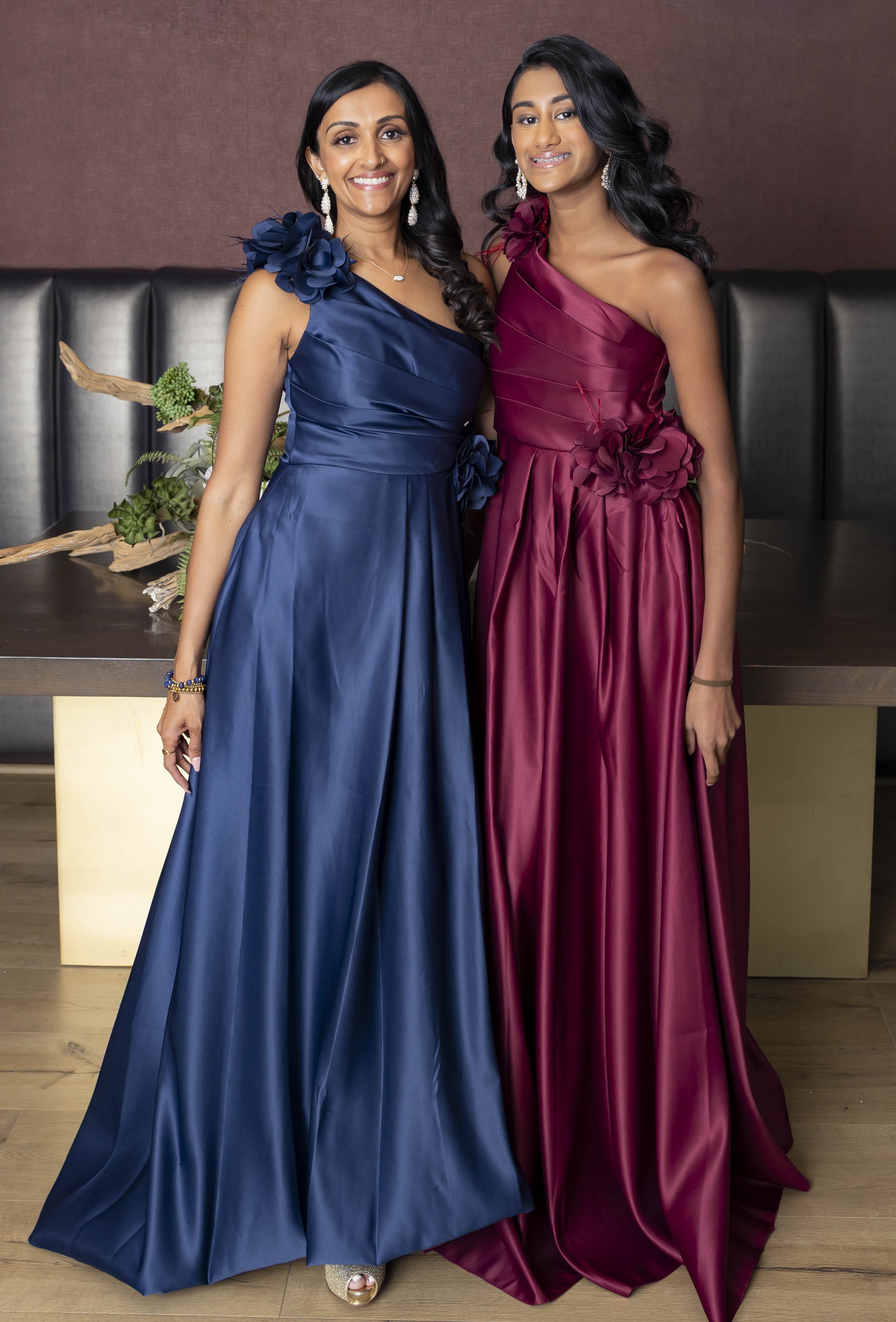 Two women in elegant satin evening gowns, one in navy blue with a shoulder flower detail and the other in deep red with floral accents at the waist, standing together indoors against a brown wall and black leather seating.