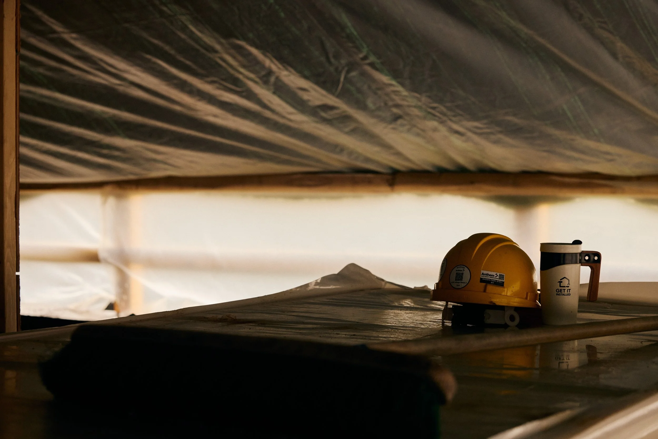 A construction helmet, a coffee mug, and a small tool on a wooden surface under a roof, with construction materials visible in the background.