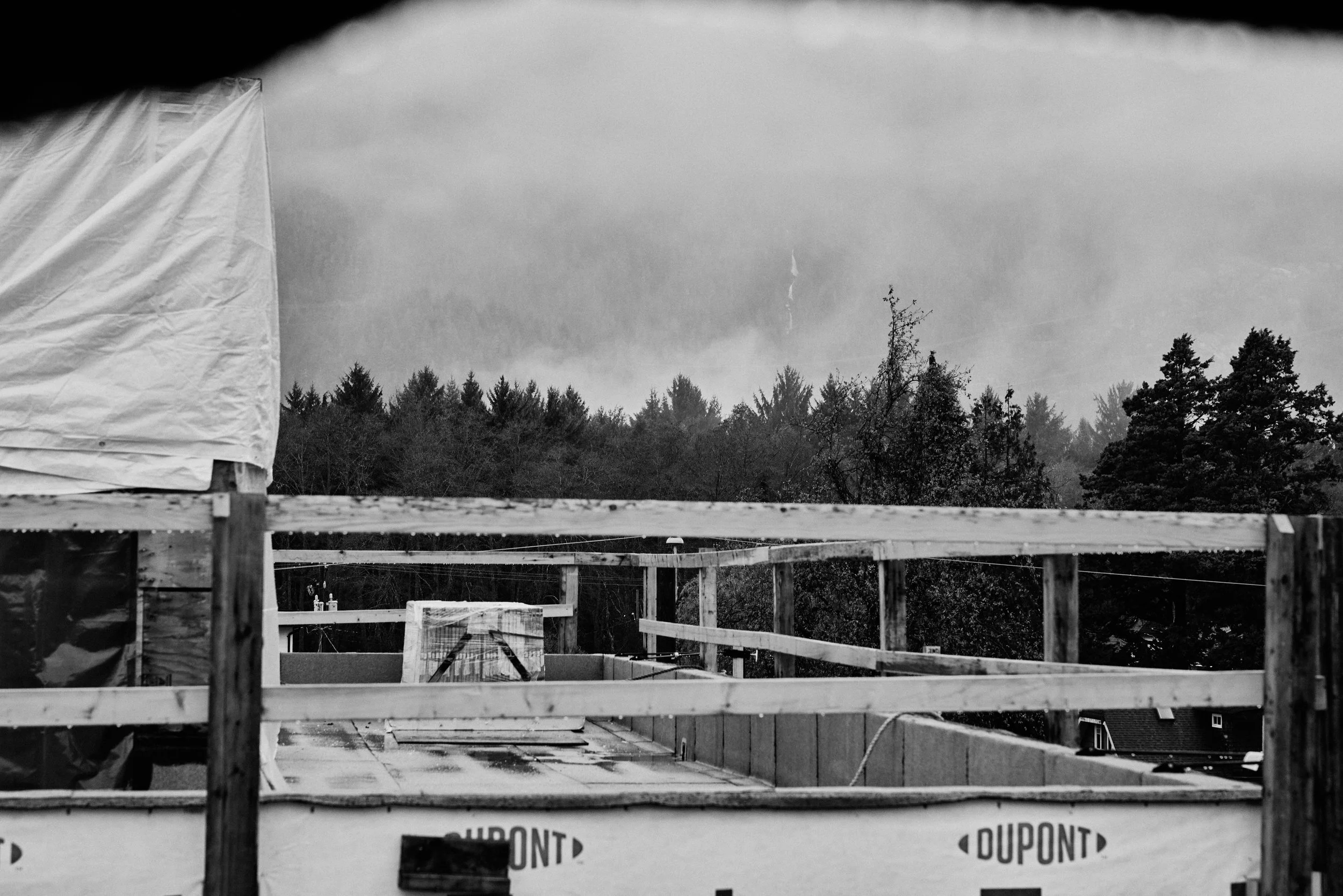 Construction site with wooden framing and partial plastic covering, set against a backdrop of trees and a cloudy sky.