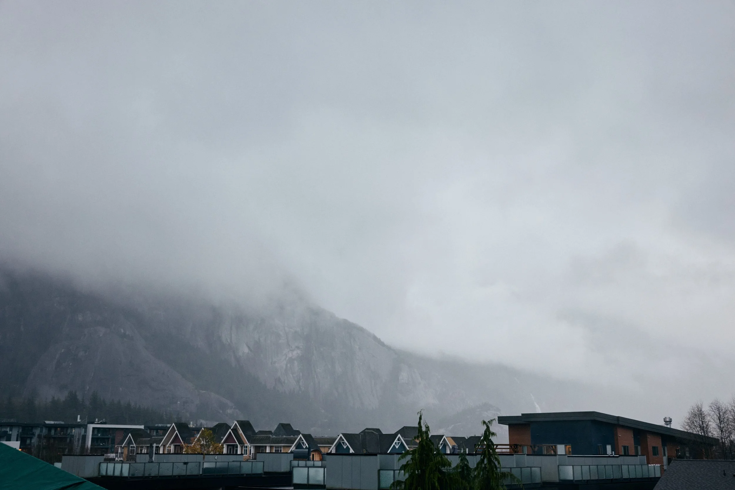 Overcast sky with low-hanging clouds over mountains in the background and rooftops of buildings in a town or city in the foreground.