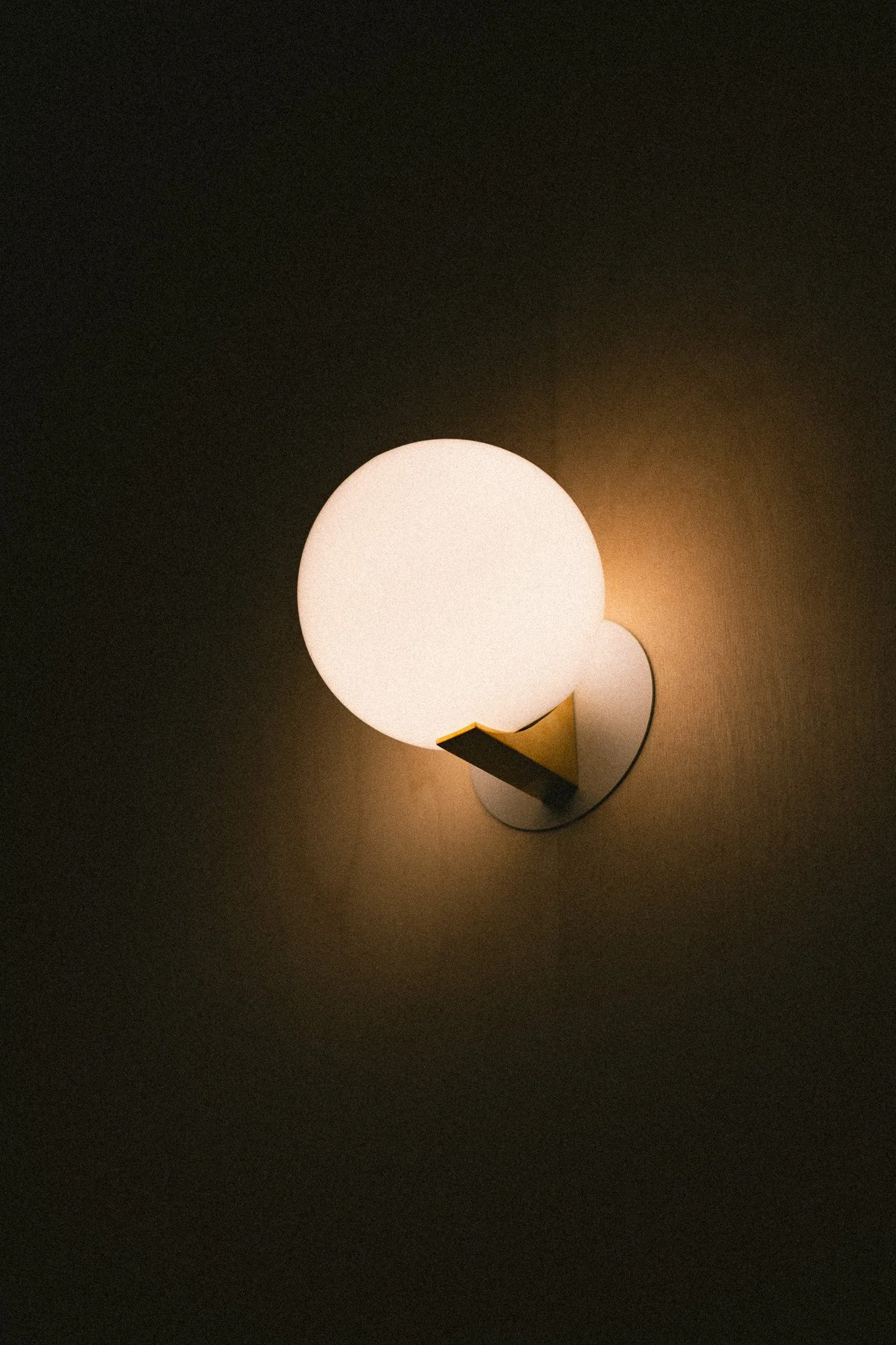 Wall-mounted modern lamp with a spherical white light and a triangular yellow accent, casting a soft glow on the wall.