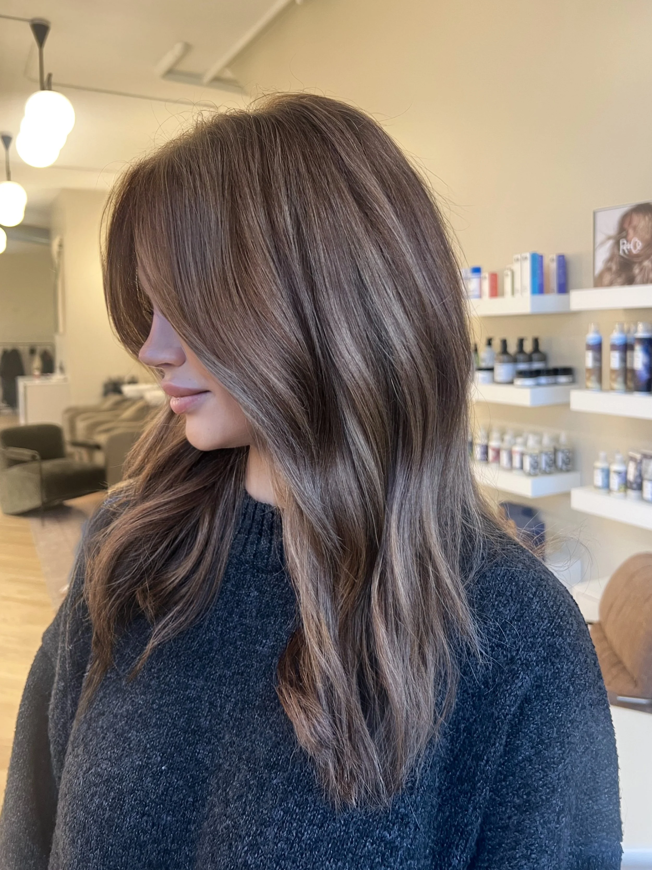 Woman with shoulder-length brown hair styled in loose waves, wearing a black top, in a salon with shelves of hair care products in the background, her hair was done by Monica Renee a blonde specialist in the Chicago area.