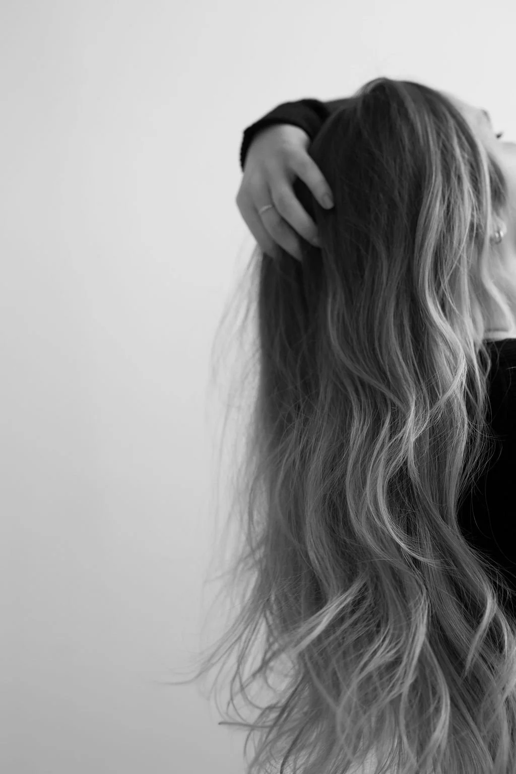 A person with long, wavy hair raising their hand to their head, partially covering their face, in black and white.