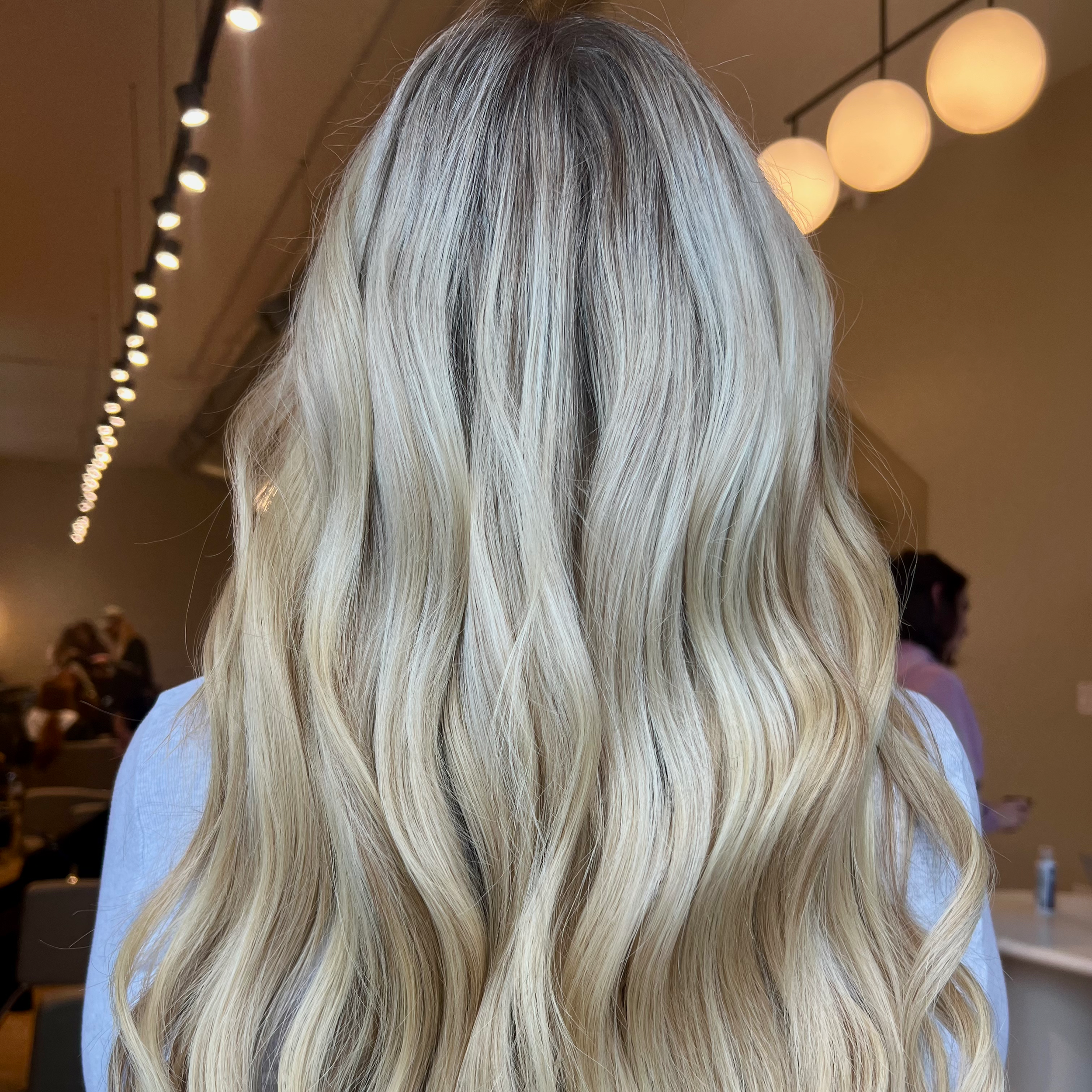 Back of a woman's head with long, wavy, icy blonde hair, colored by Monica Renee in Chicago, in a salon with round hanging lights and other people in the background.
