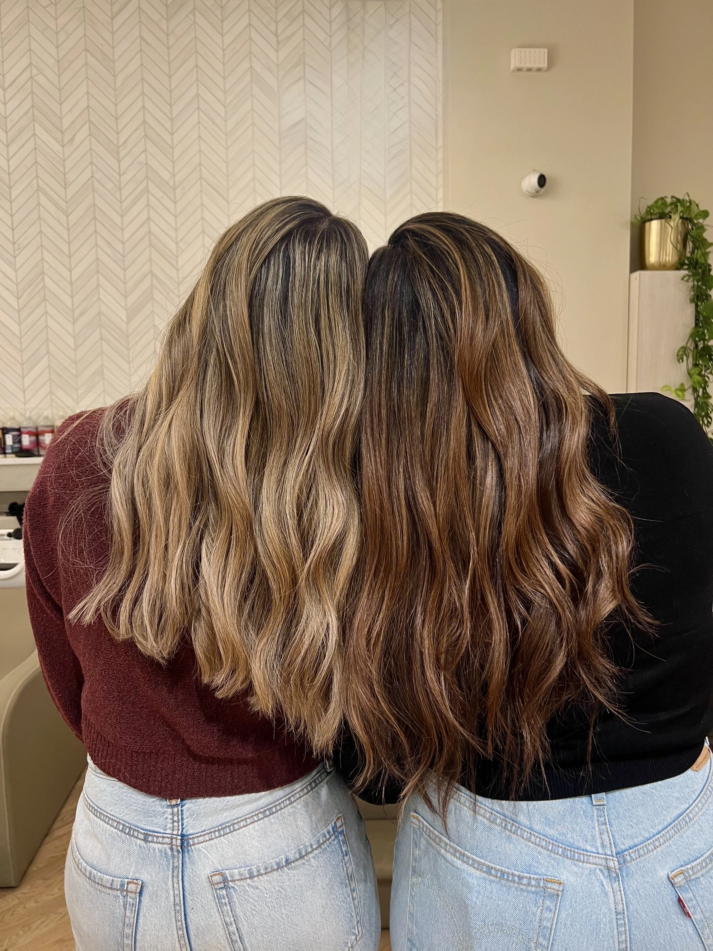 Two women with long, wavy hair, one with ash blonde highlights and the other with brunette highlights, standing close together with their backs facing the camera in a room with neutral decor. Both had their color and styles done by Monica Renee.