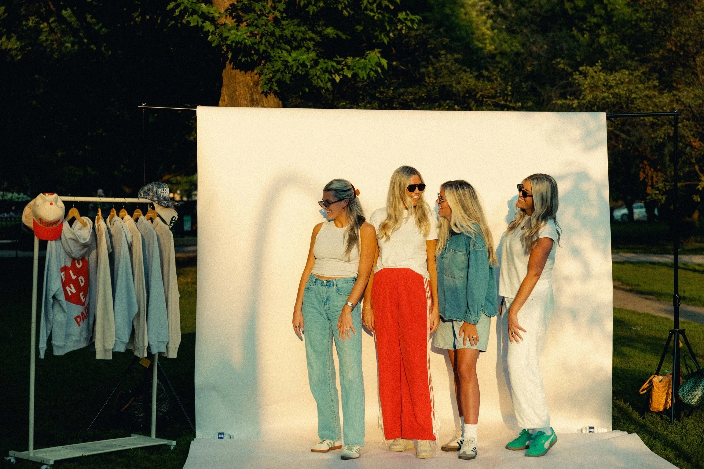 Four blonde women, styled by Monica Renee blonde specialist in Chicago, standing and smiling on a white photo backdrop outdoors during sunset, with clothing racks and hats to the left.
