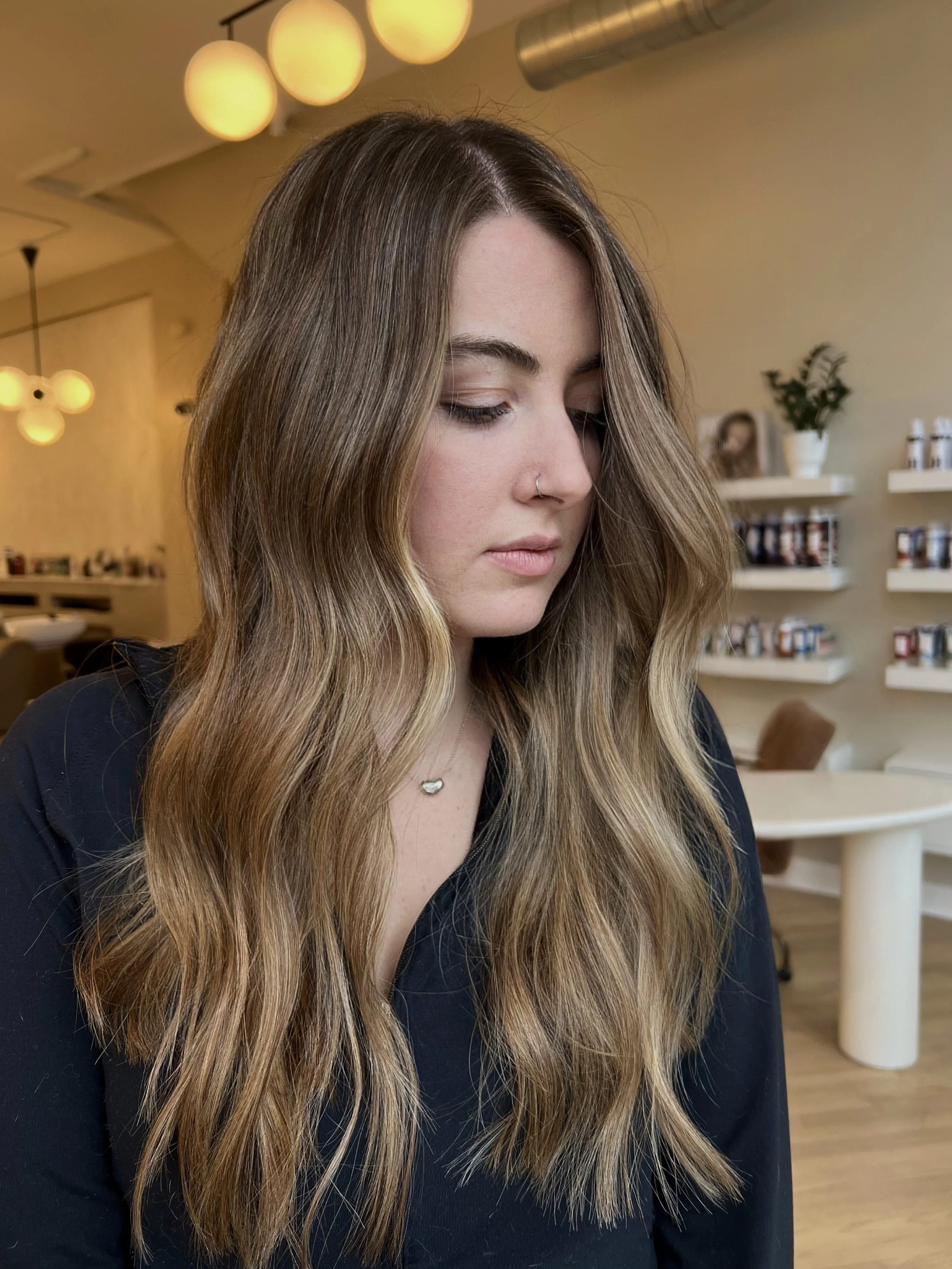 A young woman with wavy, light brown hair with blonde highlights, wearing a dark top and a small necklace, looking down with eyes closed.  Her hair was  colored and cut by Monica Renee a celebrity and influencer hairstylist in Chicago.