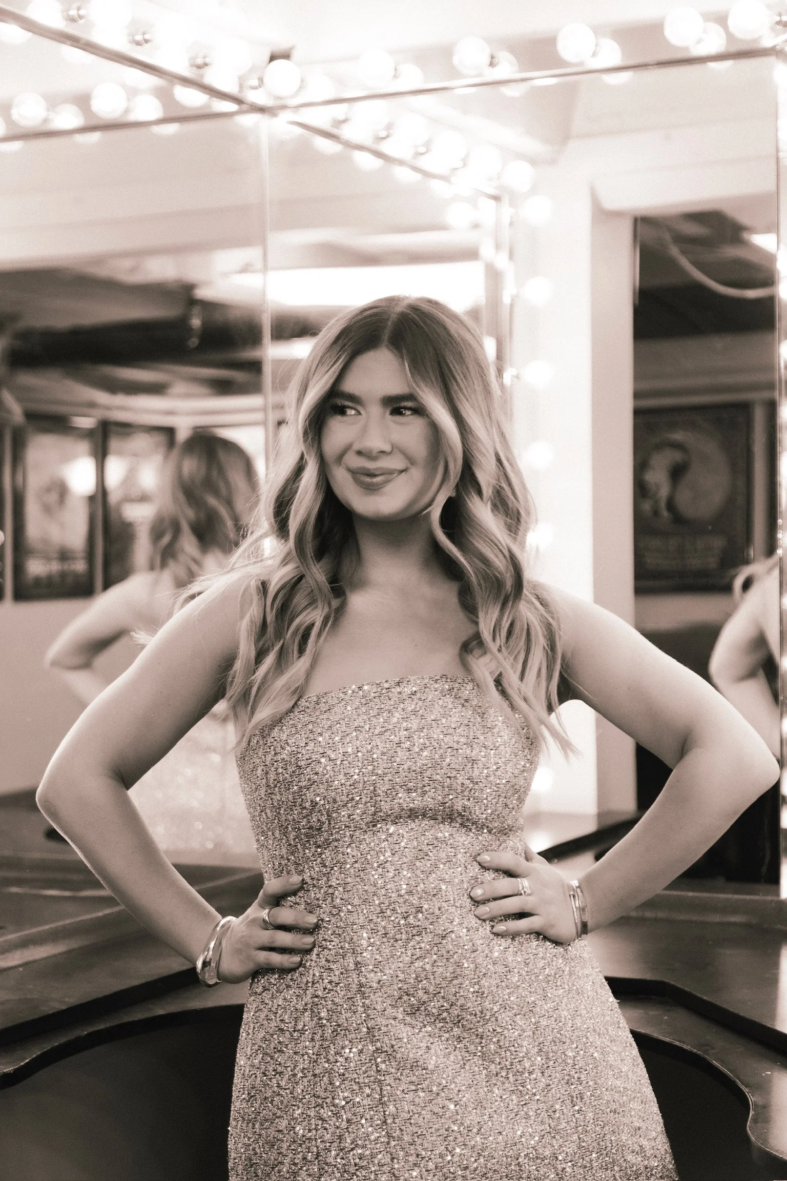 A picture of Danielle Carolan and influencer and podcaster, wearing a strapless, glittery dress, with her hair styled by Monica Renee who is a celebrity hairstylist in Chicago, standing in front of a mirror with bulbs around it.