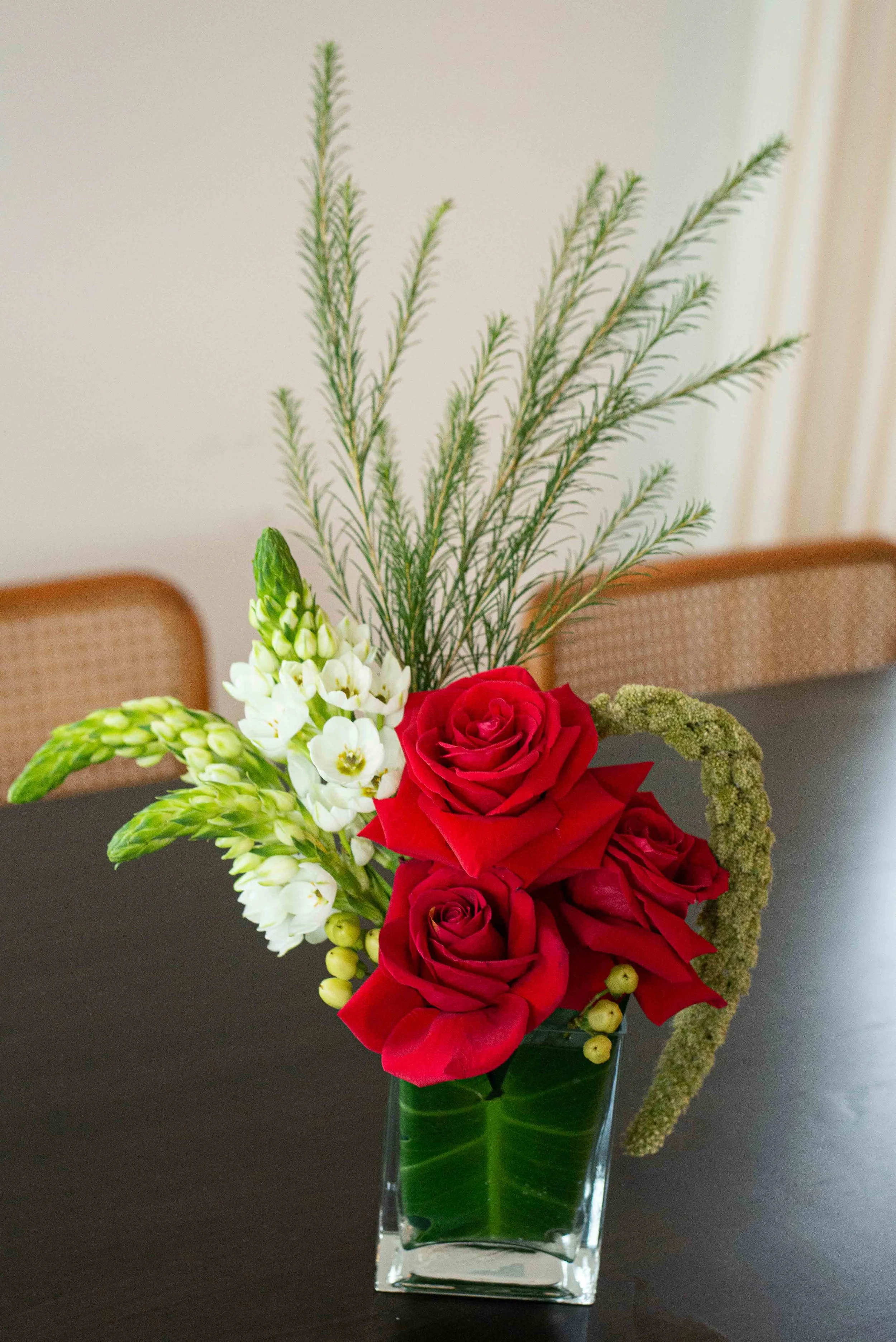 A floral arrangement in a clear square glass vase containing red roses, white flowers, green foliage, and a tall sprig of rosemary on a black table with a wooden chair and beige wall in the background.