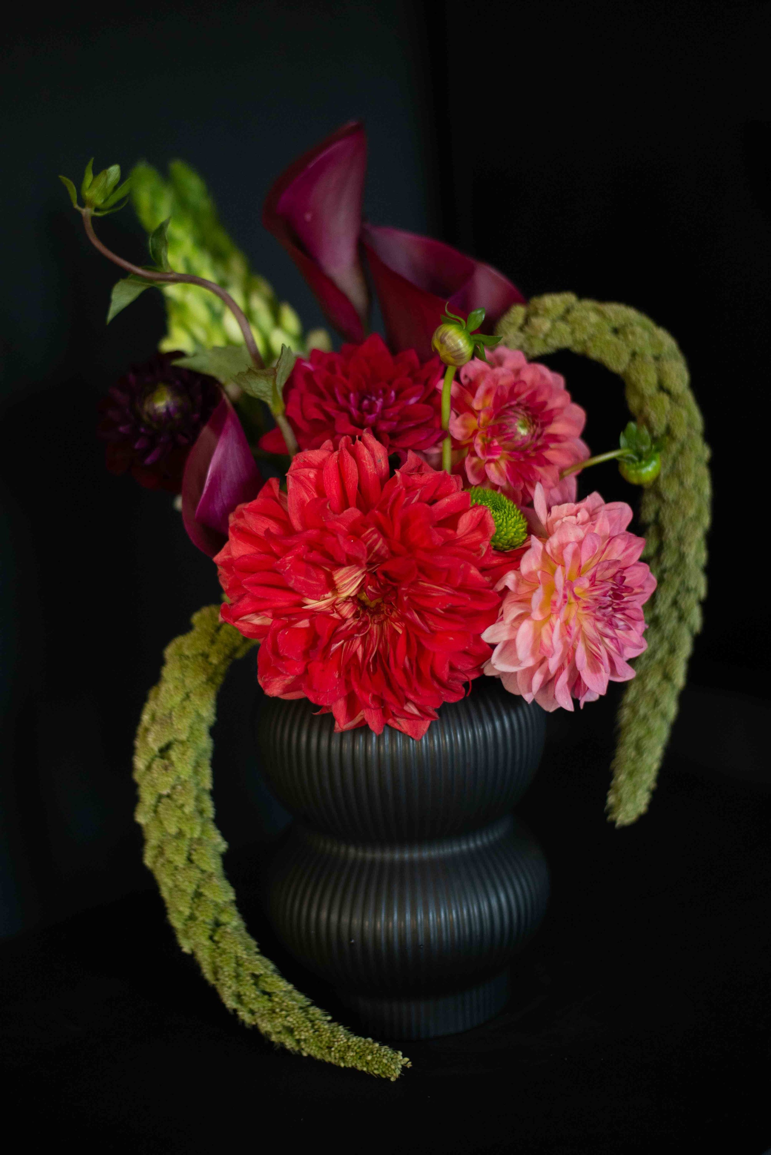 A black vase with ridges holds a colorful arrangement of flowers including red and pink dahlias, calla lilies, green leaves, and trailing green amaranthus, set against a black background.