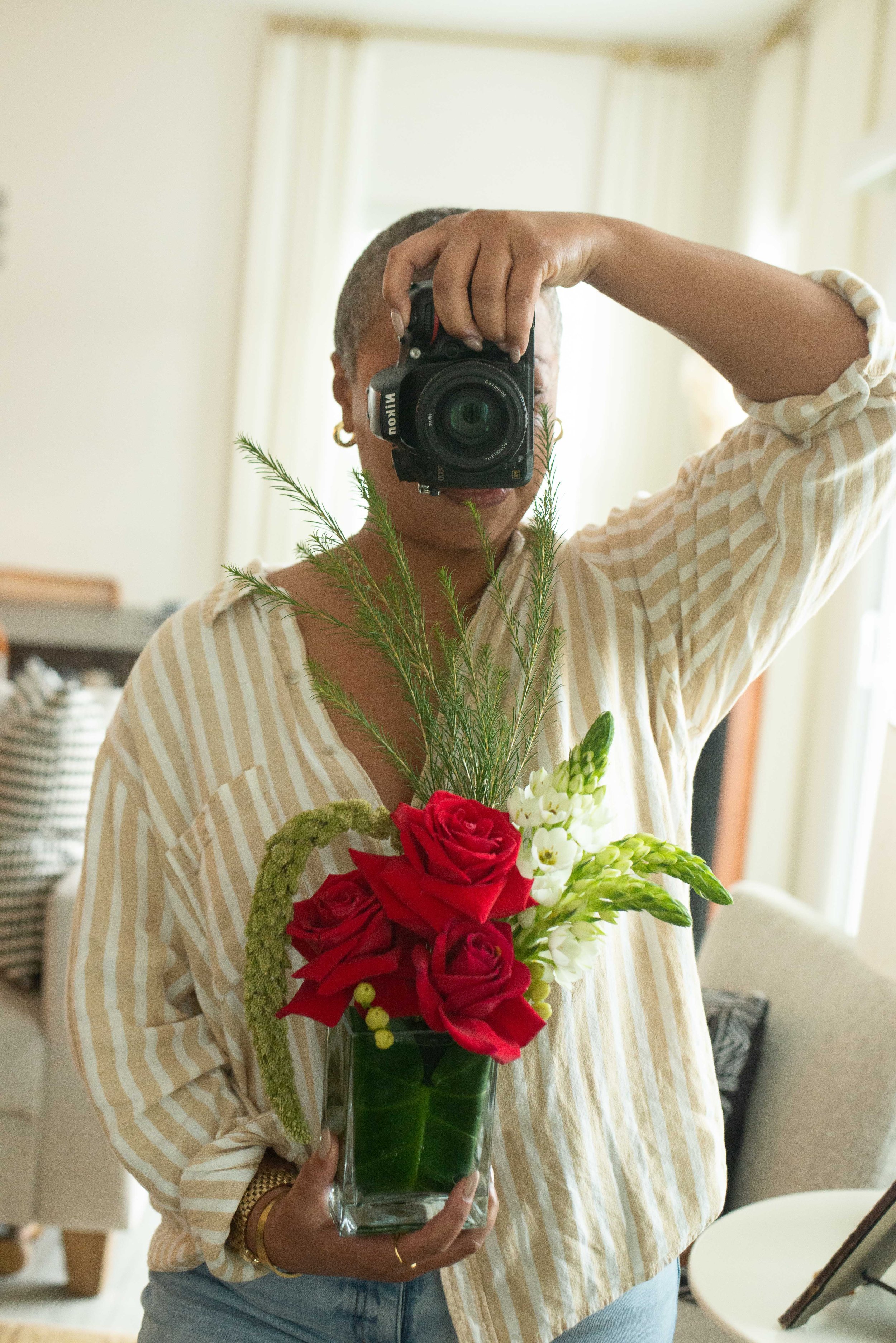 Person taking a mirror selfie holding a flower arrangement with red roses, white flowers, and green foliage in a glass vase, wearing a light striped shirt and wrist jewelry.