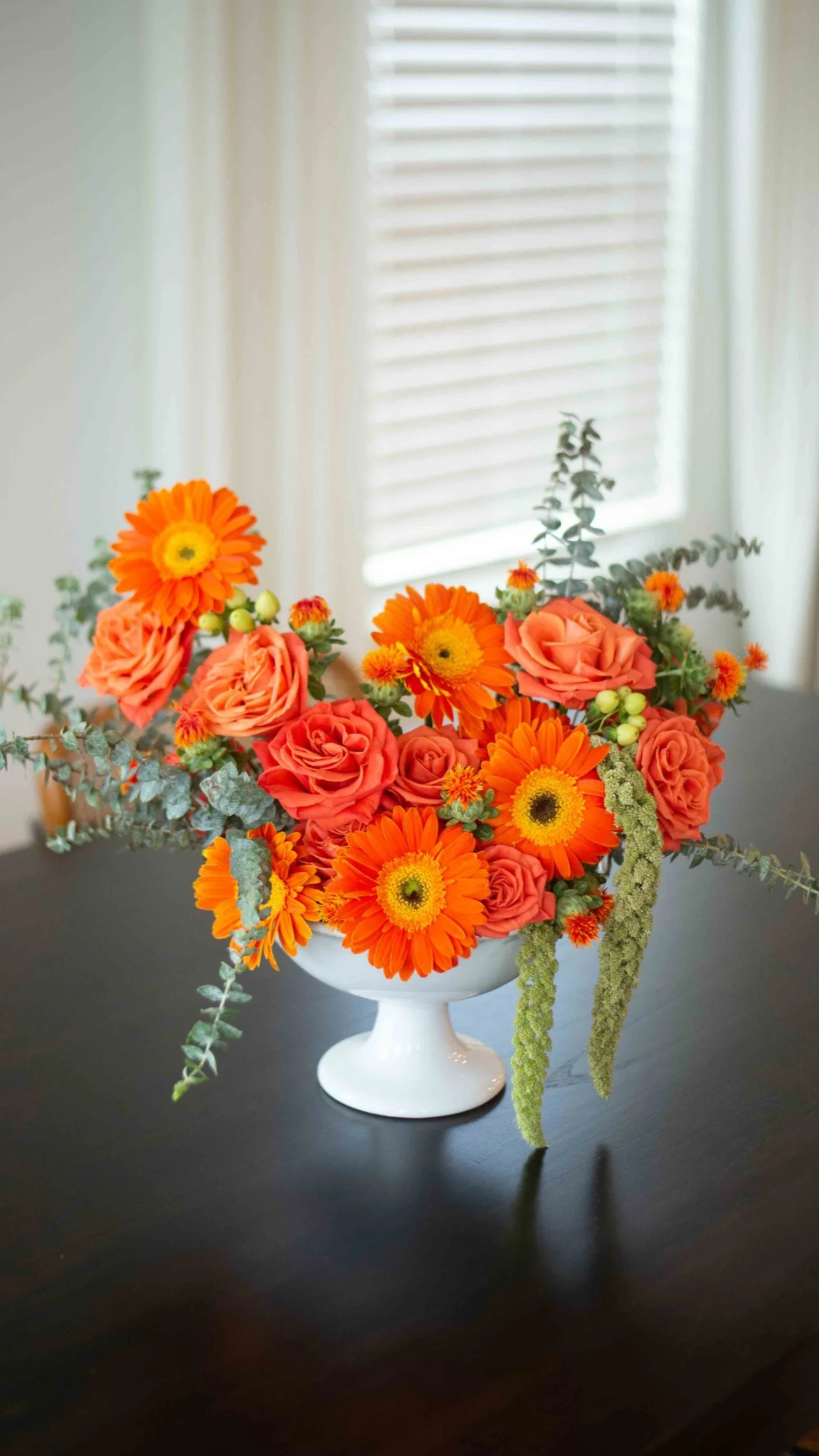 charlotte, boutique florist , A bouquet of orange and peach roses and orange gerbera daisies in a white pedestal vase, with gray-green foliage, placed on a dark table near a window with white blinds.