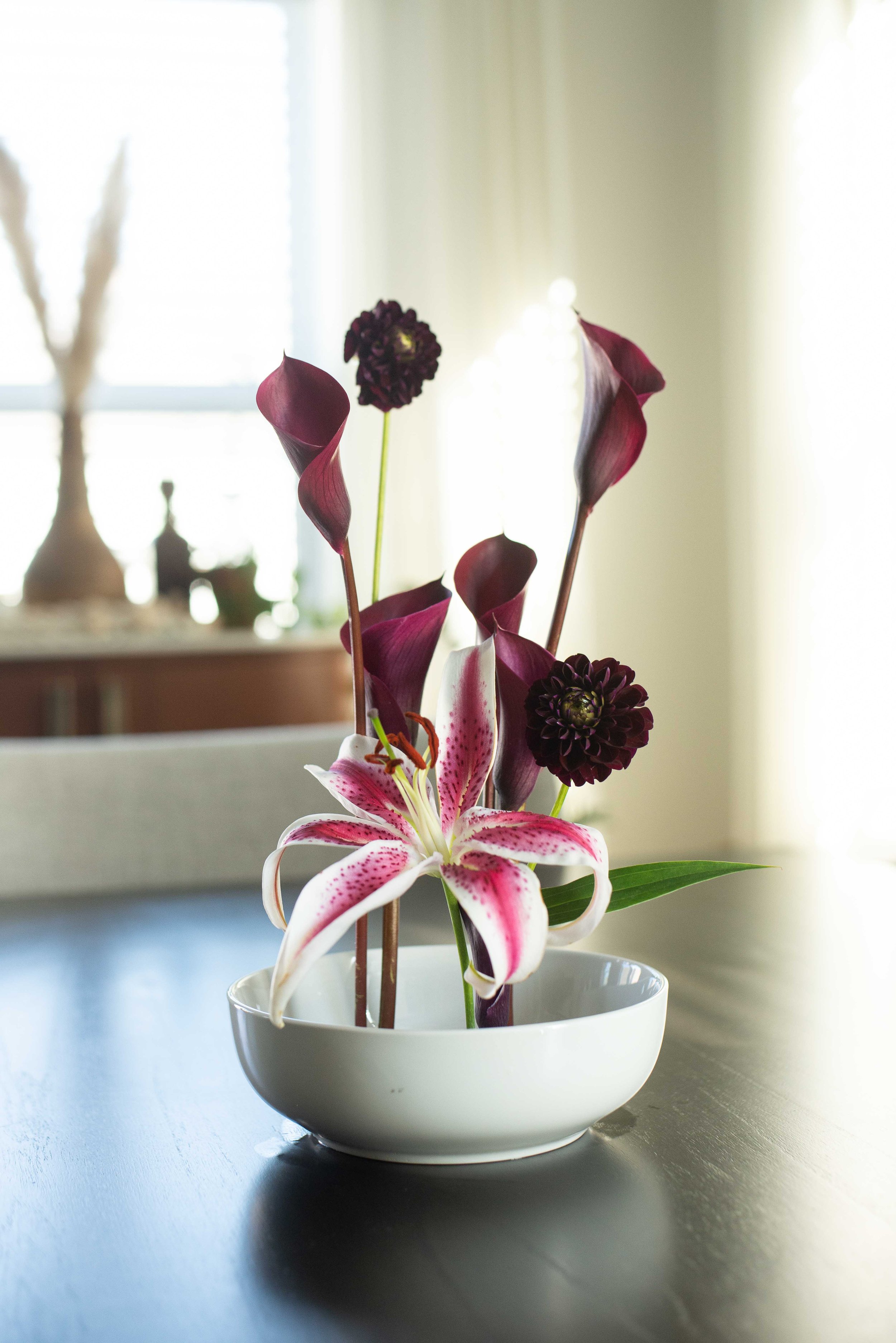 A floral arrangement in a white bowl featuring pink and white lily flowers and dark purple calla lilies, with a blurred background of a room with window light.