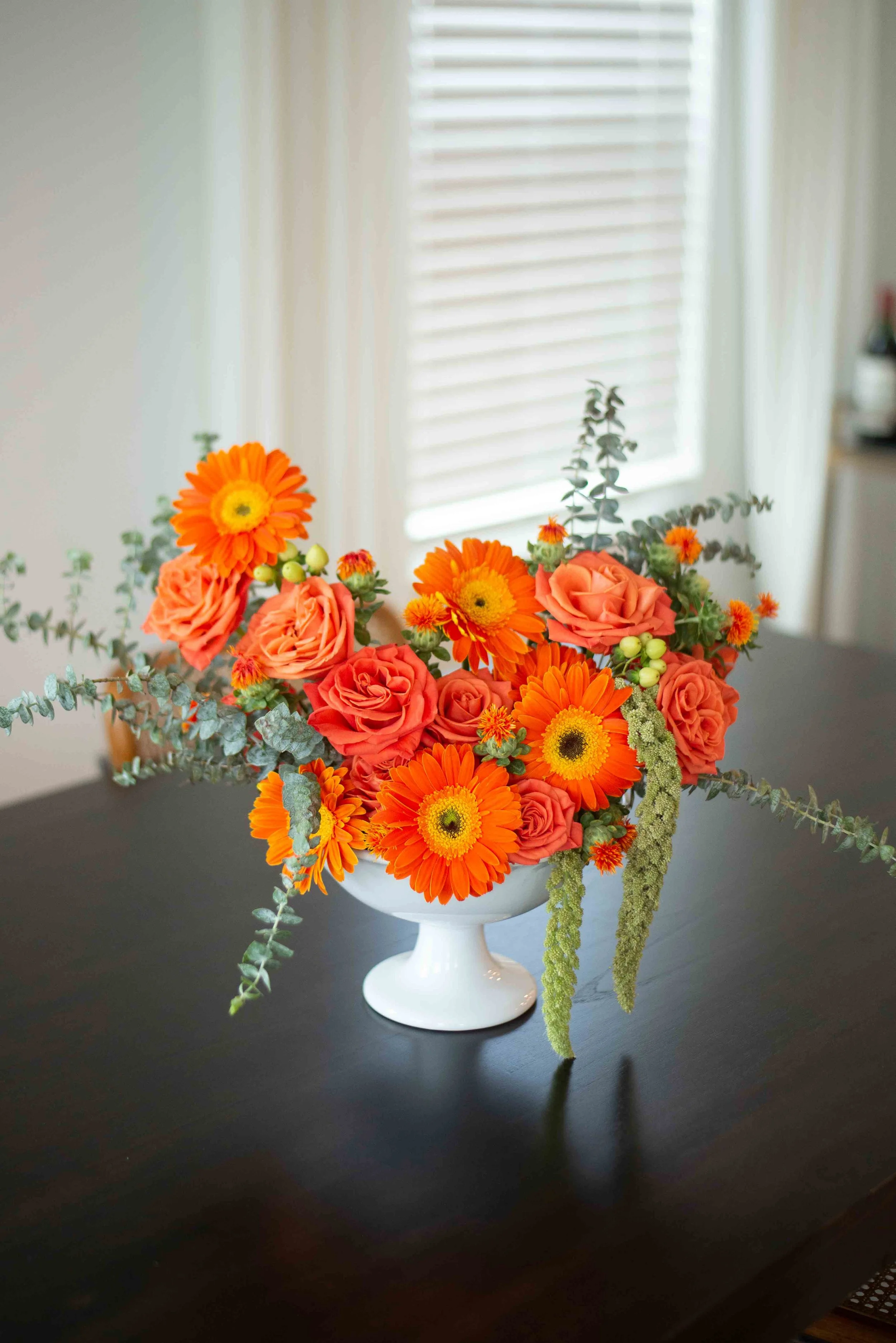 A bouquet of orange roses, orange gerbera daisies, and green foliage in a white pedestal vase on a black table, with a window with white blinds in the background.