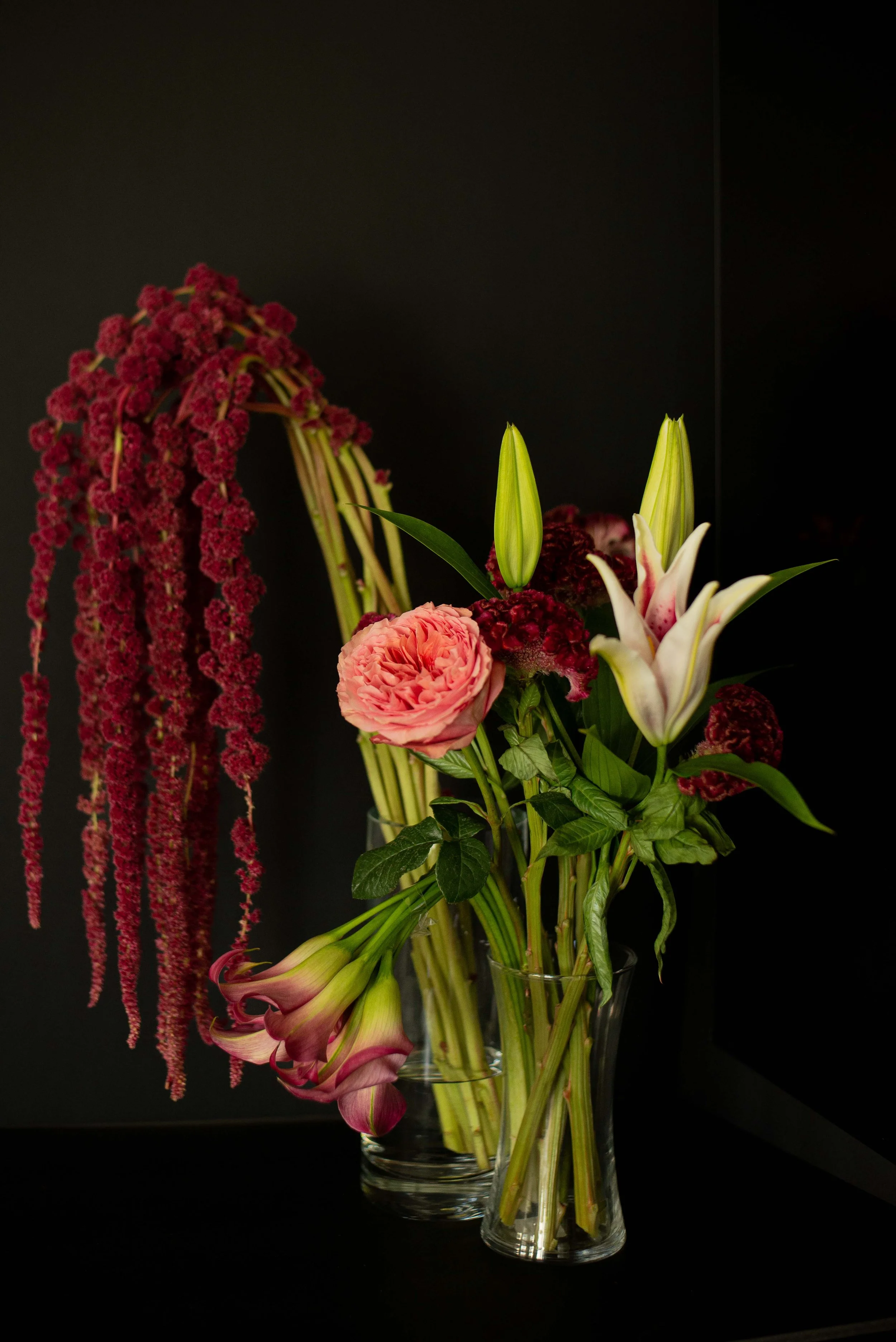 A glass vase holding a bouquet of pink, white, and red flowers, including lilies and a ranunculus, against a black backdrop.