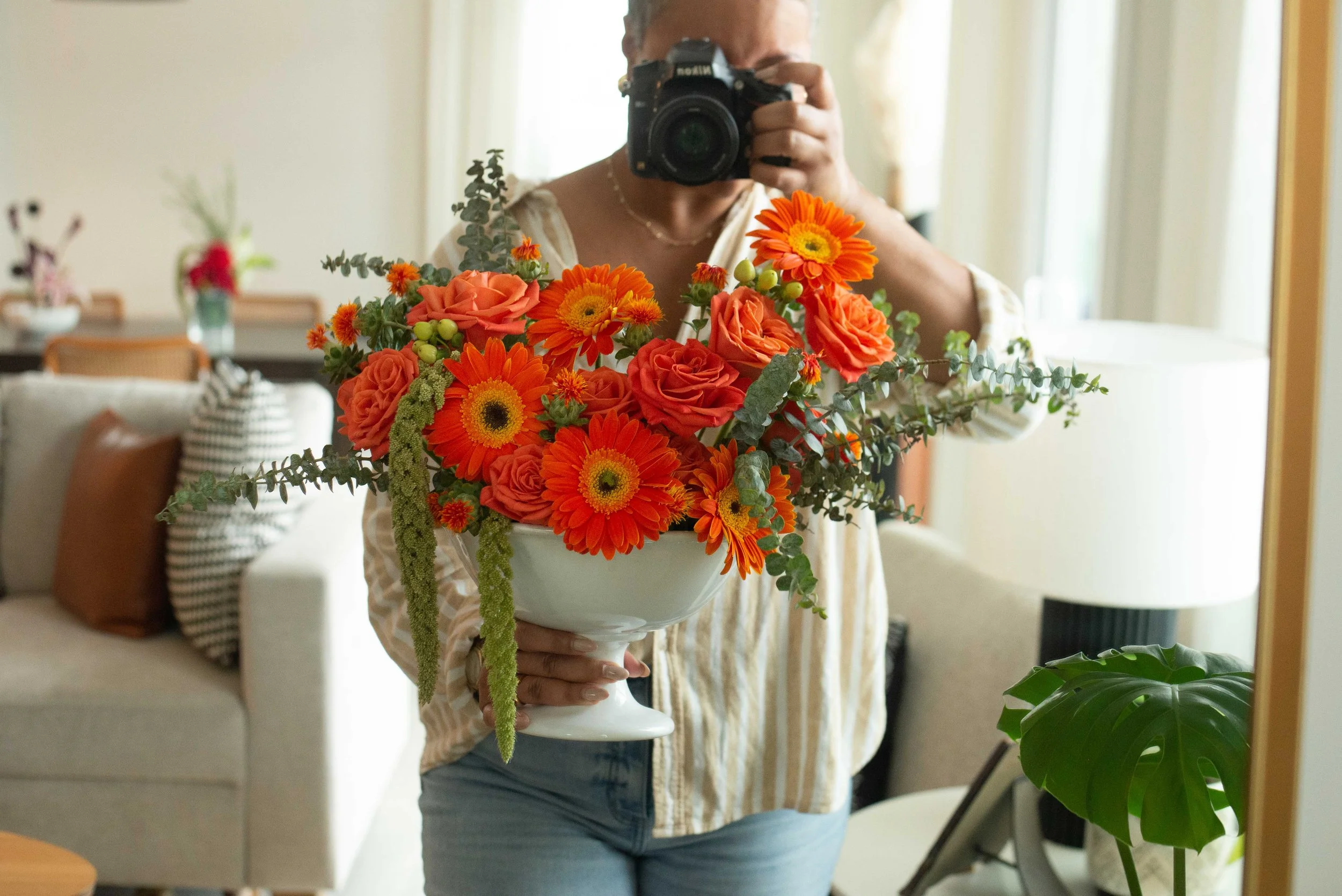 Person taking a mirror selfie with a camera while holding a white vase of orange and coral flowers in a bright living room.