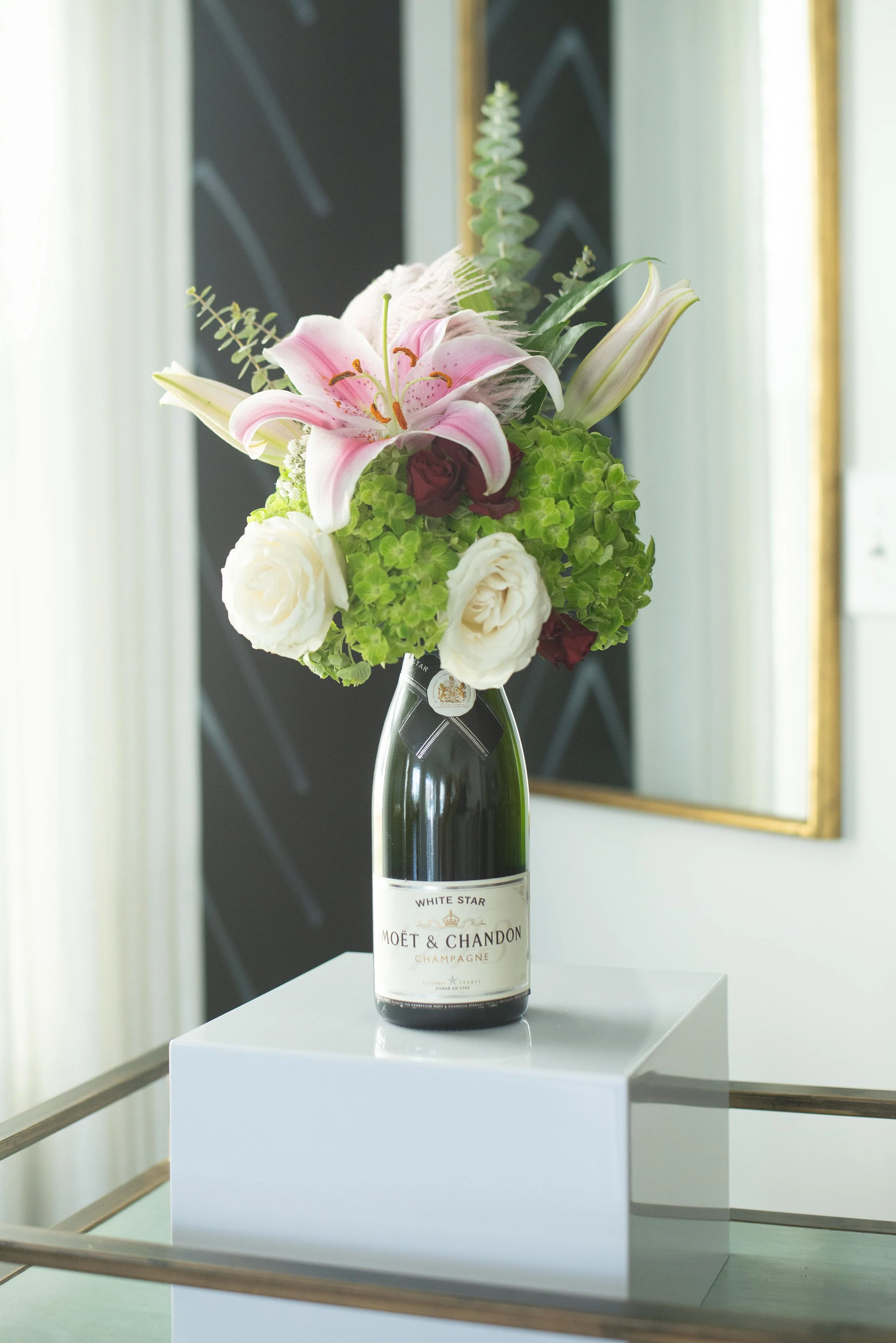 A flower arrangement in a Moët & Chandon champagne bottle on a white pedestal in a gallery setting.