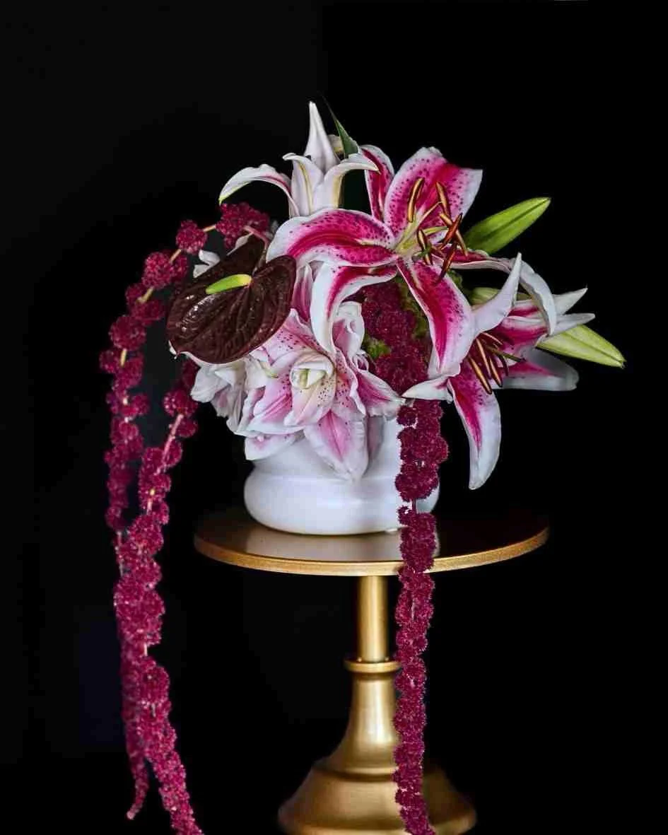 Arrangement of pink and white lilies, dark purple leaves, and pink stringy flowers in a white vase on a gold stand against a black background.
