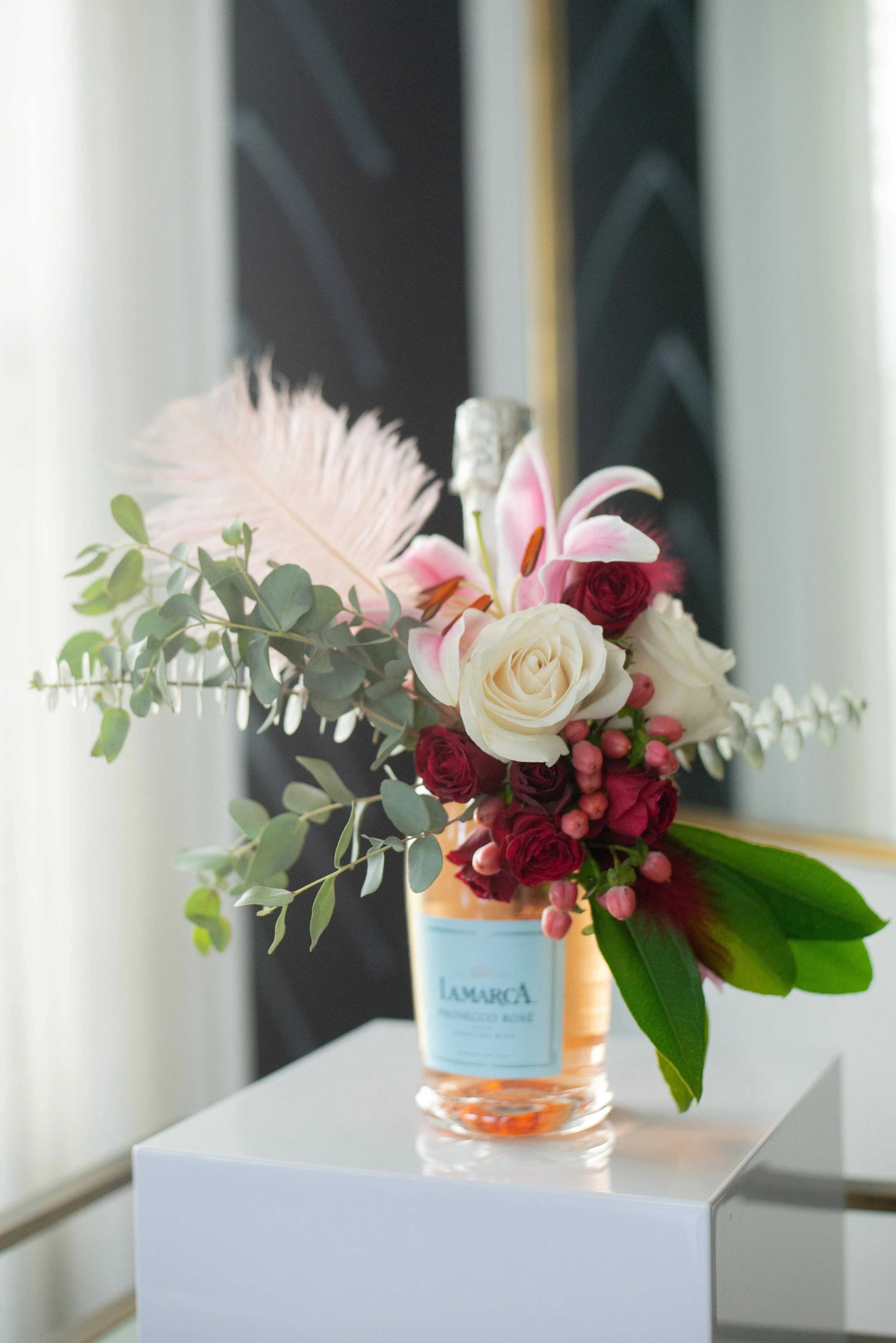 Floral bottle styling experience by Moe, A bouquet of flowers in a vase on a white table, with a blurred blackboard or dark wall in the background.