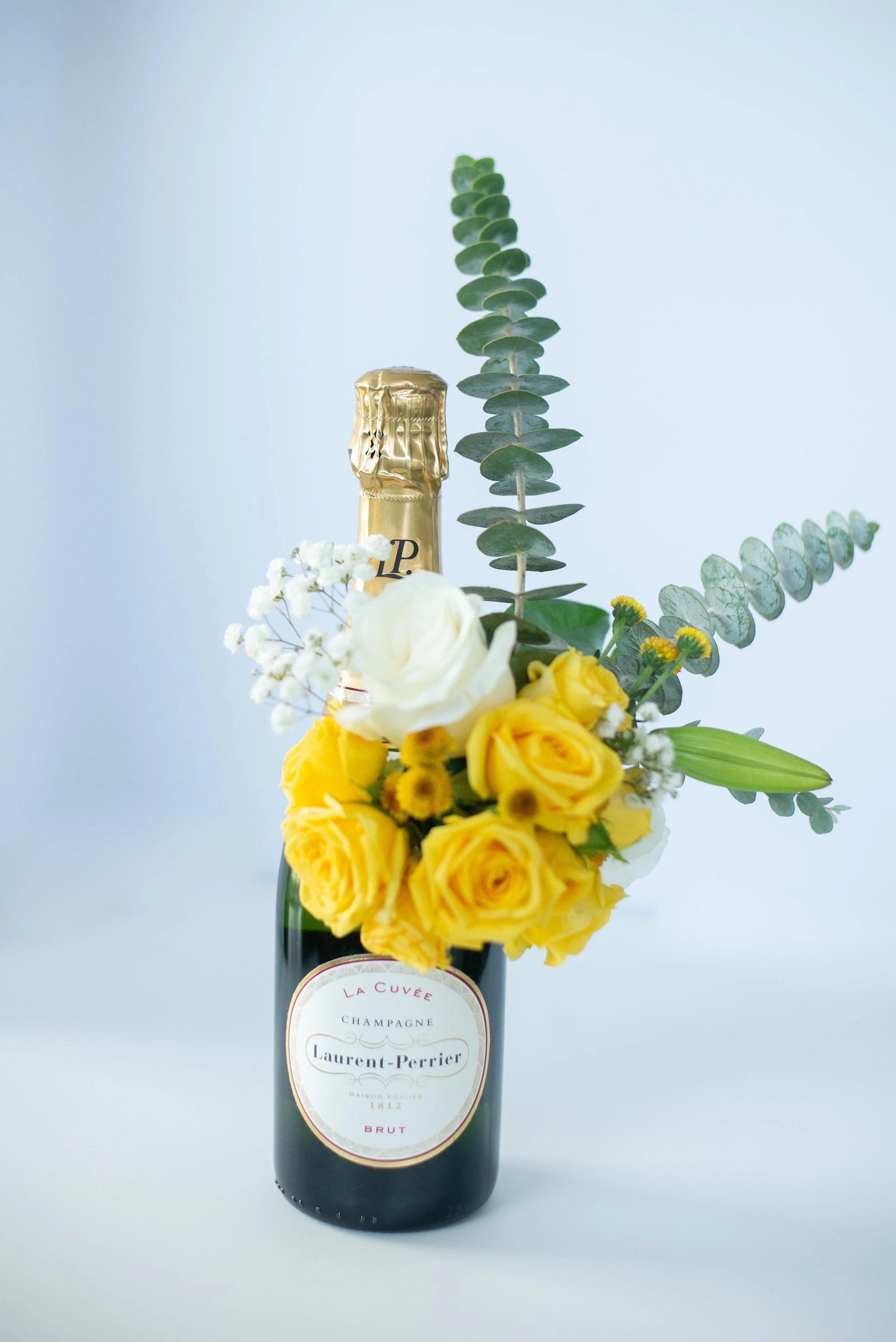 A bottle of Laurent-Perrier champagne decorated with a floral arrangement of yellow roses, white flowers, and eucalyptus leaves against a pale blue background.