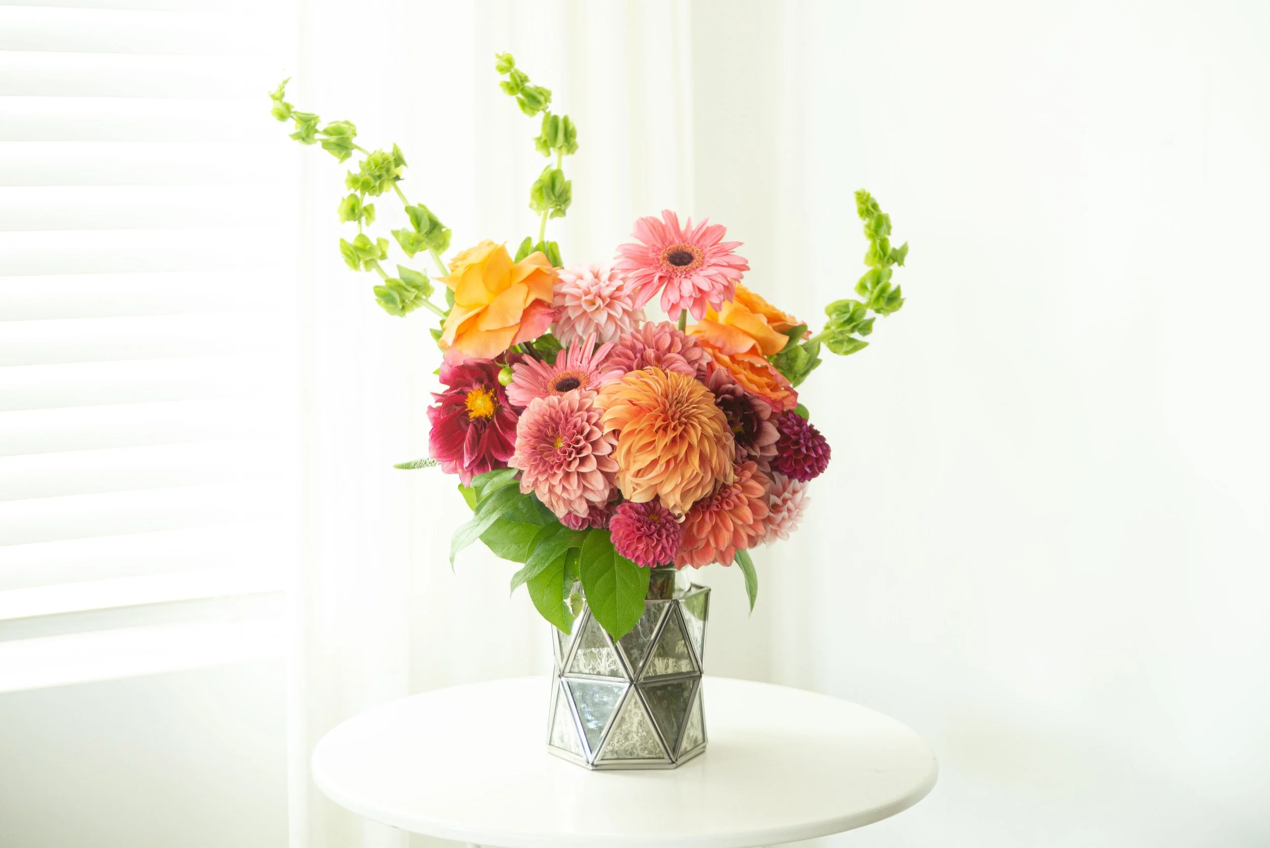 A colorful bouquet of flowers, including pink gerbera daisies and orange dahlias, arranged in a geometric glass vase on a white table, with a white background and blinds.