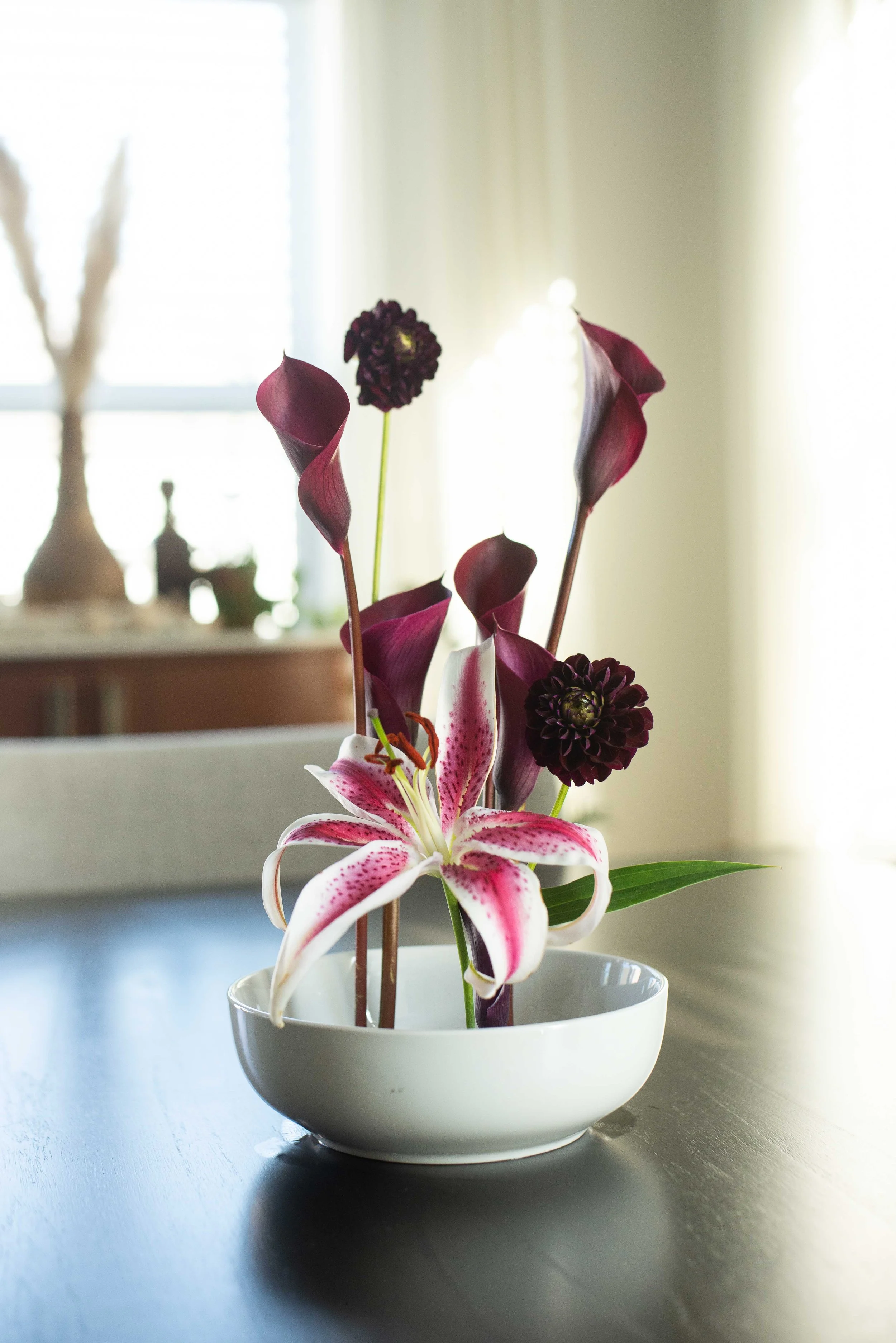 A white bowl with a floral arrangement of lilies, calla lilies, and dark dahlias on a dark wooden table, with light coming from a bright window in the background.