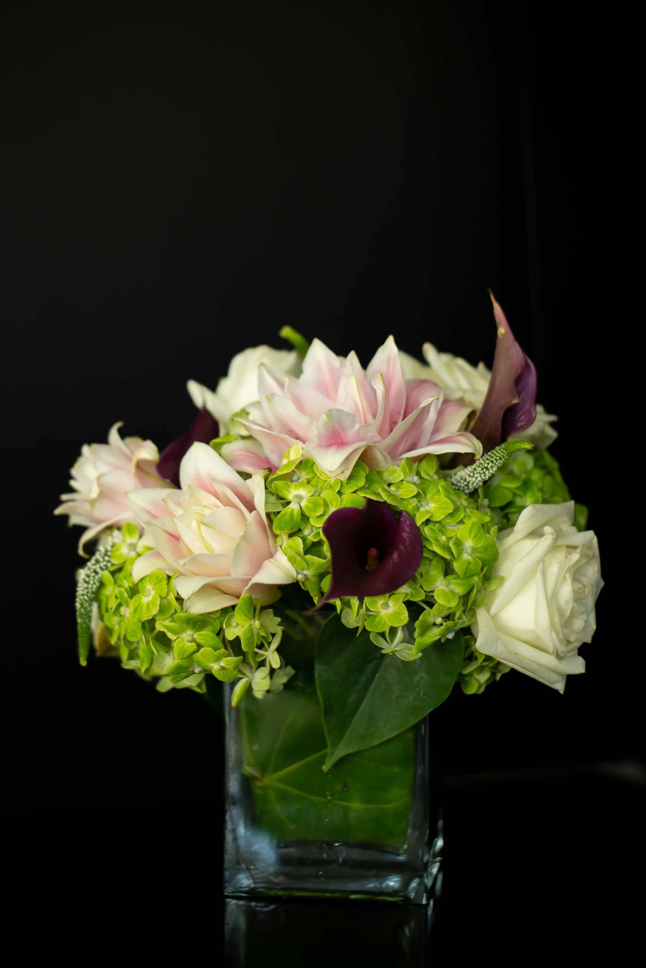 A floral arrangement with pink and white flowers, green leaves, and purple accents in a clear glass vase against a black background.