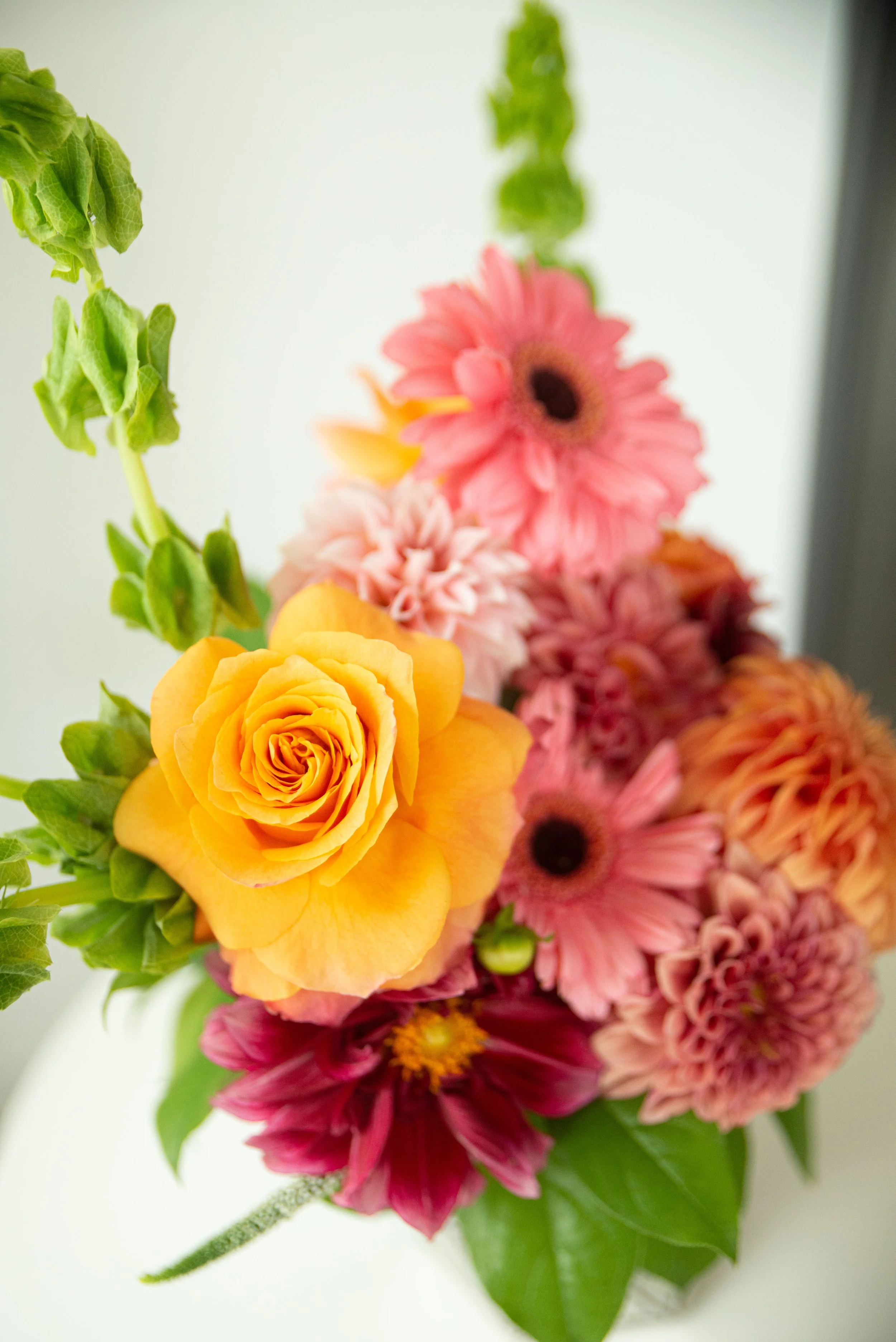 Close-up of a vibrant bouquet of flowers, including yellow roses, pink gerbera daisies, and other colorful blossoms with green foliage, against a light background.
