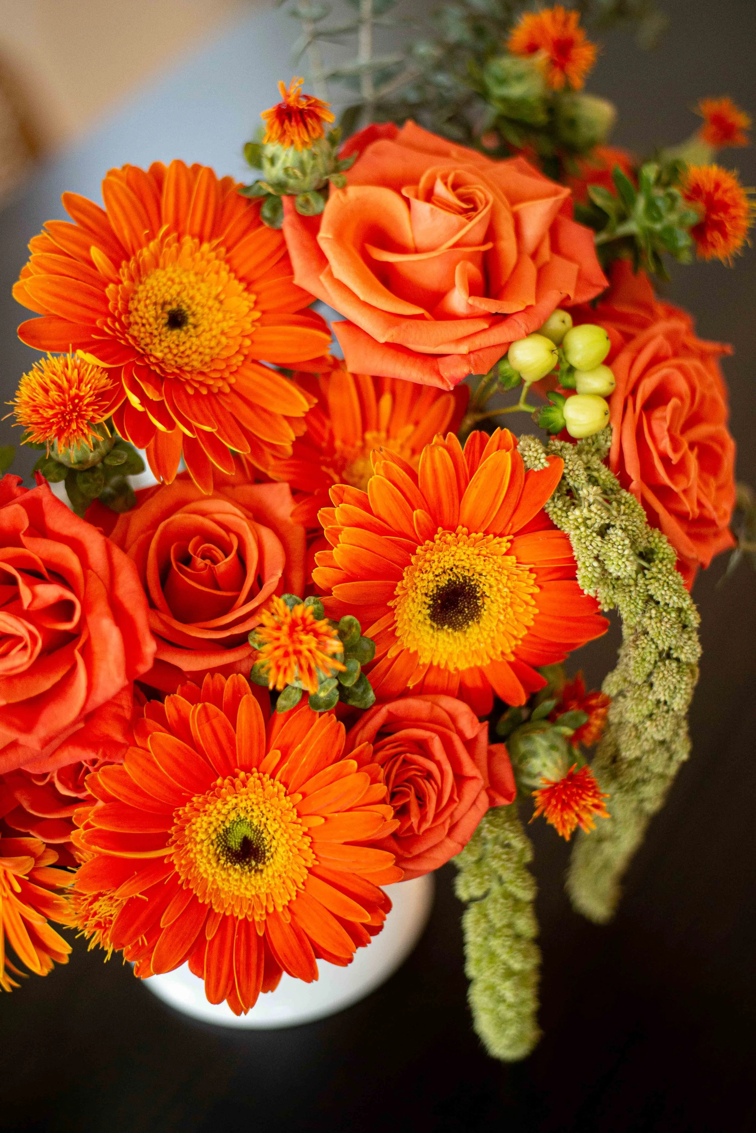 Close-up of a bouquet of orange roses, orange gerbera daisies, small orange and red flowers, and green buds and foliage, with a dark background.