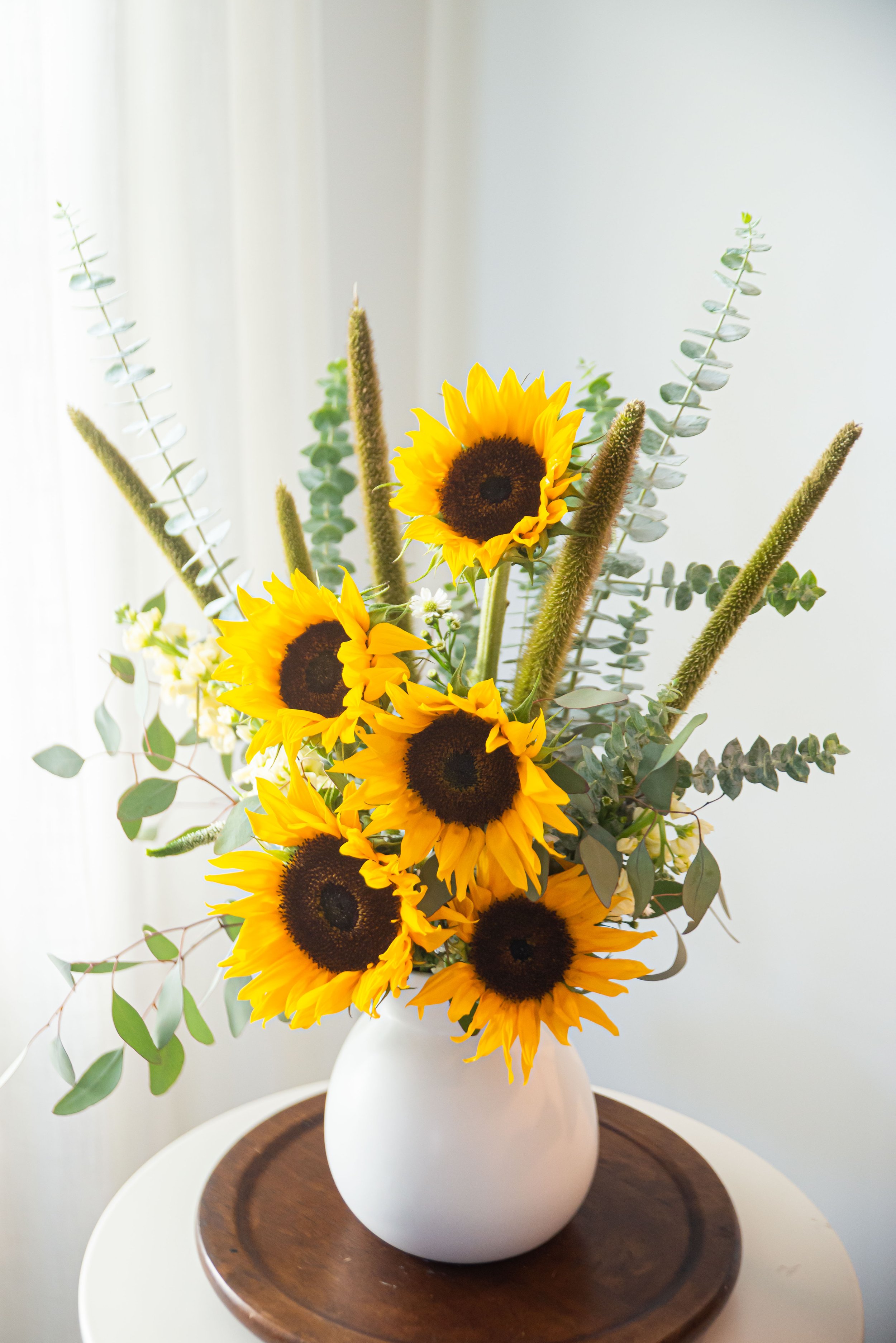 A bouquet of bright yellow sunflowers with dark centers, accented by eucalyptus leaves in a rounded white vase, placed on a wooden tray on a white side table.