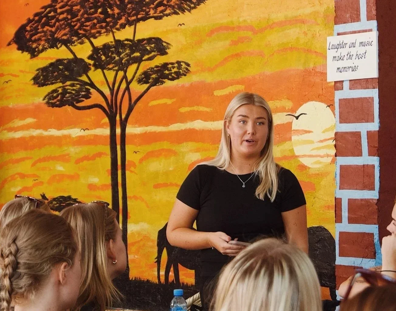 En ung kvinna håller ett föredrag för en grupp ungdomar i ett klassrum, med ett väggmålning av en soluppgång och träd i bakgrunden.