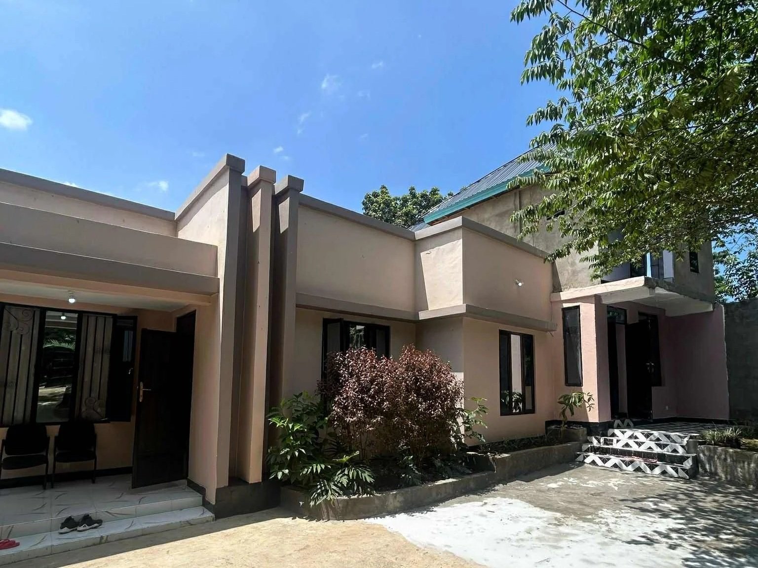 En modern tvåvåningsvilla med beige fasad, stora fönster och en liten trädgård med buskar och träd under en klar blå himmel.