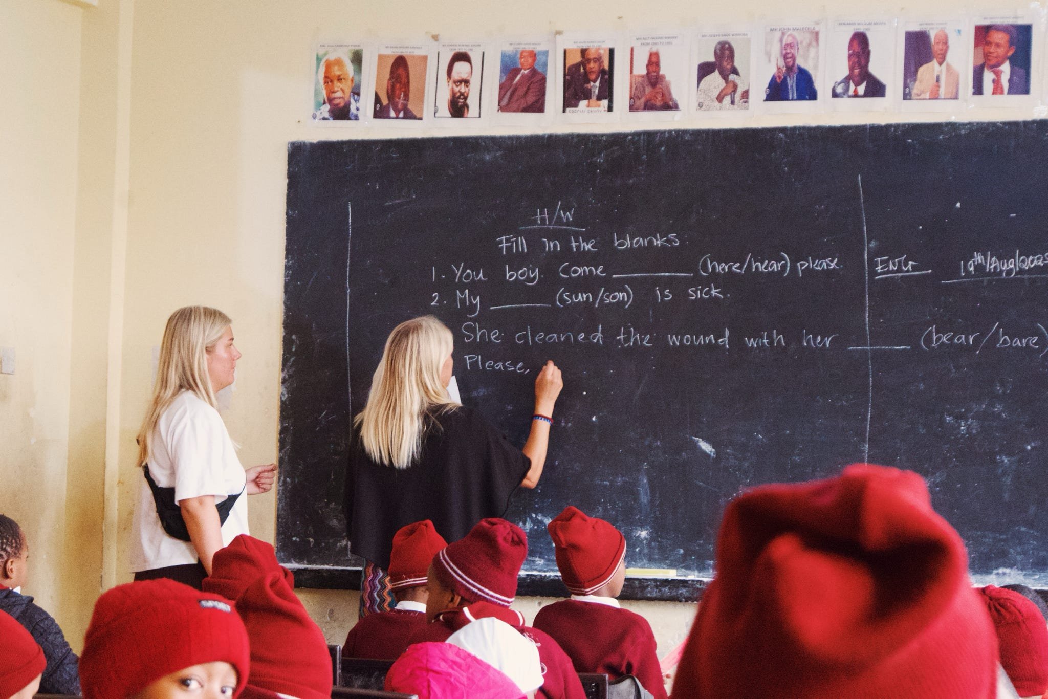 En klassrumsscen med en lärare som skriver på en svart tavla och ett barn som deltar. Flera elever bär röda mössor.