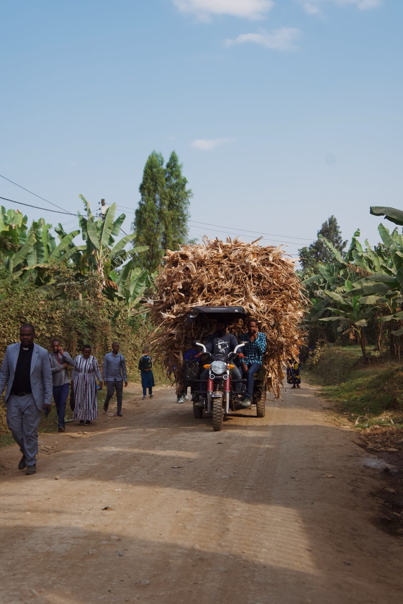 En motorcykel med ett sidovagn som är lastad med mycket torkad växtmaterial på en landsväg, omgiven av personer och gröna träd.