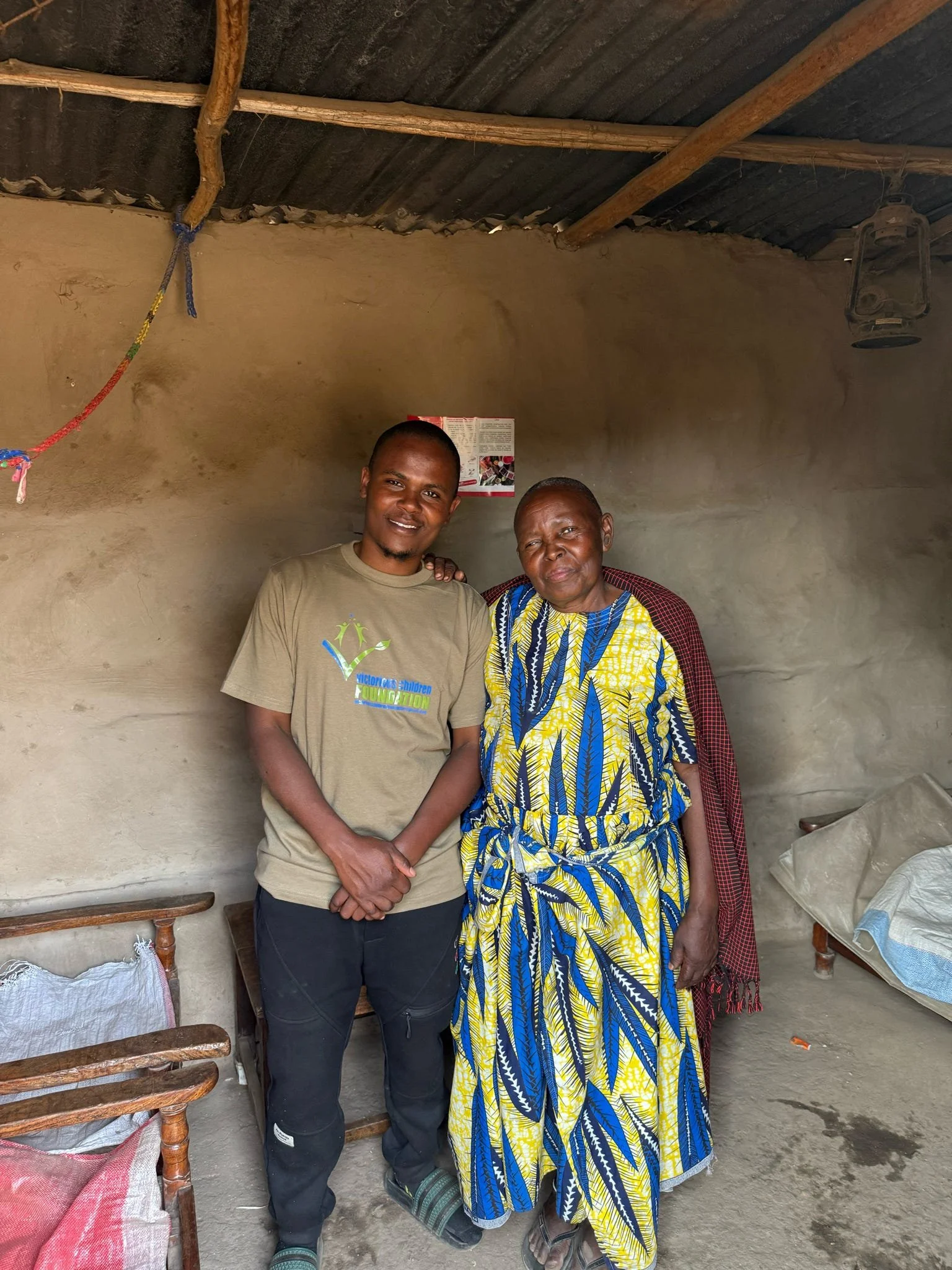 Två personer står tillsammans i ett enkelt rum med lerväggar och ett tak av trälister och corrugated plåt. Den ena personen är en ung man iklädd en beige T-shirt och svarta byxor. Den andra personen är en äldre kvinna iförd en färgglad klänning med blå, gula och svarta mönster, samt en röd schal över axlarna. De står nära varandra och tittar mot kameran.
