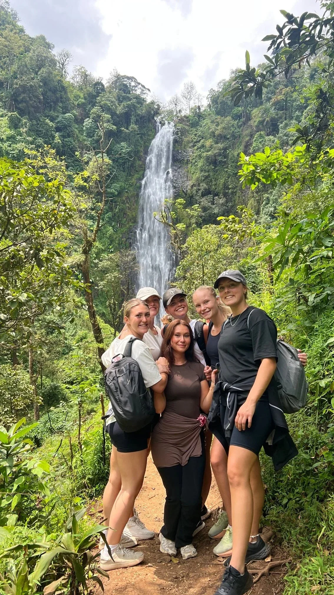 En grupp kvinnor i vandraskläder står tillsammans framför en stor vattenfall i en grön skog, med ett molnigt himmelsskydd ovanför.